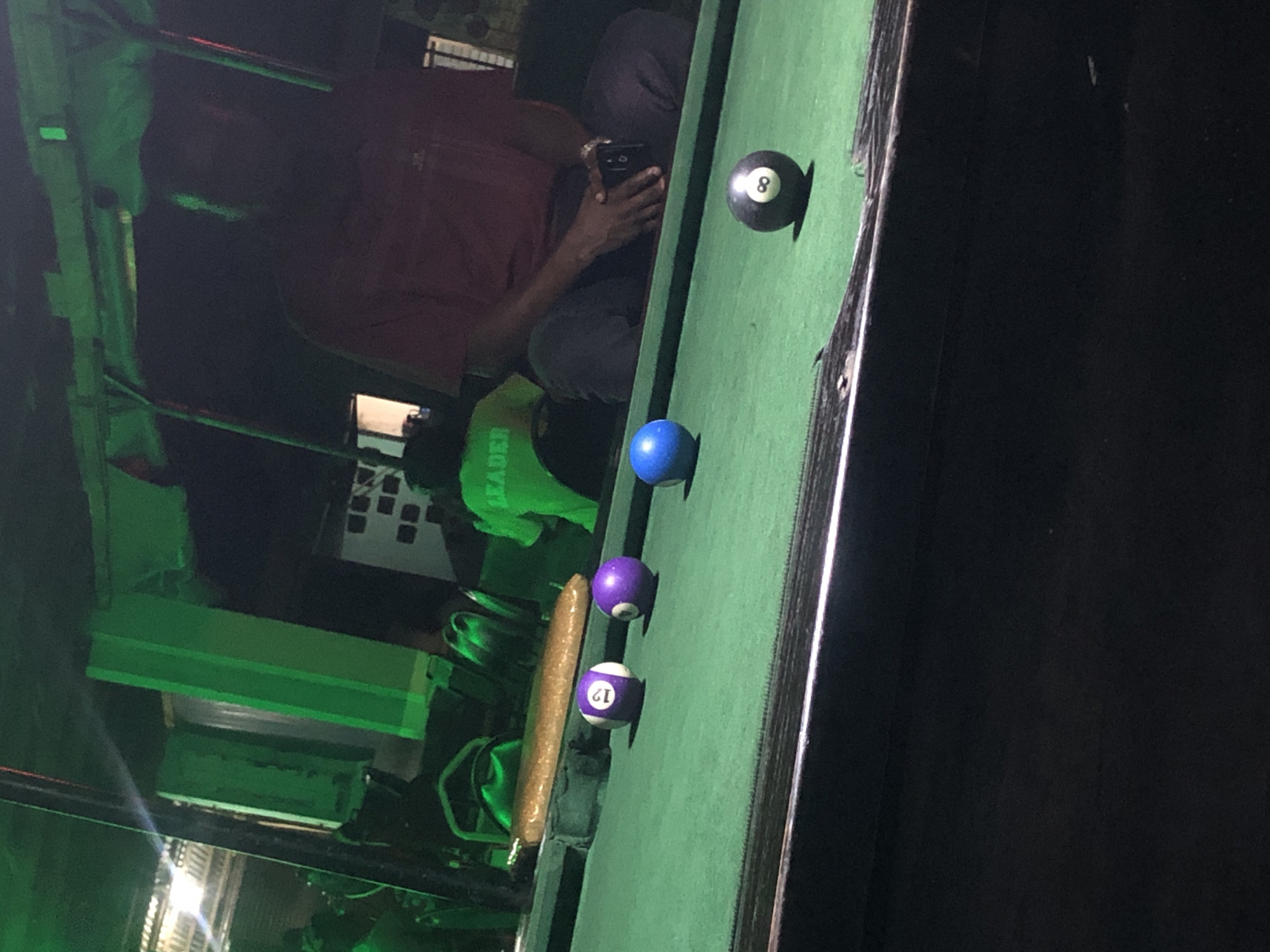 The image depicts a dimly lit indoor setting, likely a bar or recreational area, with an illuminated green pool table as the central focus. The pool table has several balls on it: a purple solid ball, a purple ball with the number 12 in white, a blue solid ball, and a black 8-ball with a white circle around the number 8. A long, golden-brown object, possibly a cue stick or a rolled-up mat, rests on the edge of the table. In the background, a person wearing a red shirt and dark pants is seated, holding a phone, with their hands clasped. Another person in a bright green shirt with the word "LEADER" visible on the back is also in the background, facing away from the camera. The overall ambiance is dark, with green lighting casting a glow on the scene. The location is identified as Unknown, Unknown.