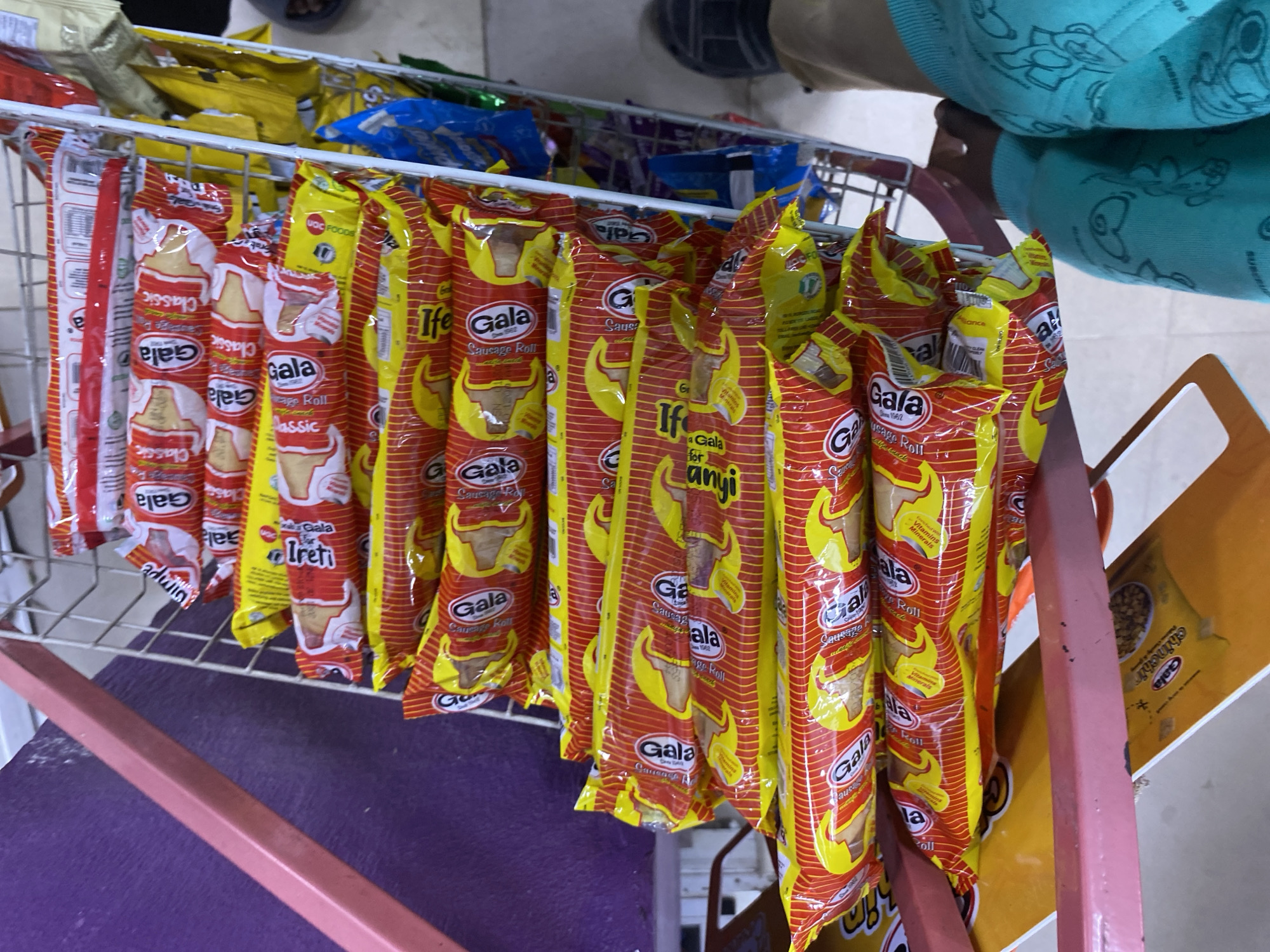 The image shows a wire rack in what appears to be a small shop or kiosk in Lagos, Nigeria. The rack is filled with various snacks, prominently featuring Gala sausage rolls in red and yellow packaging, along with other snack products. A person wearing a turquoise shirt is partially visible in the upper right corner, and their feet can be seen at the top of the image. The background is somewhat blurry, but the presence of a shelf with other packaged food items suggests a retail environment. The rack itself has a pink frame and sits on a purple surface. The scene is brightly lit.