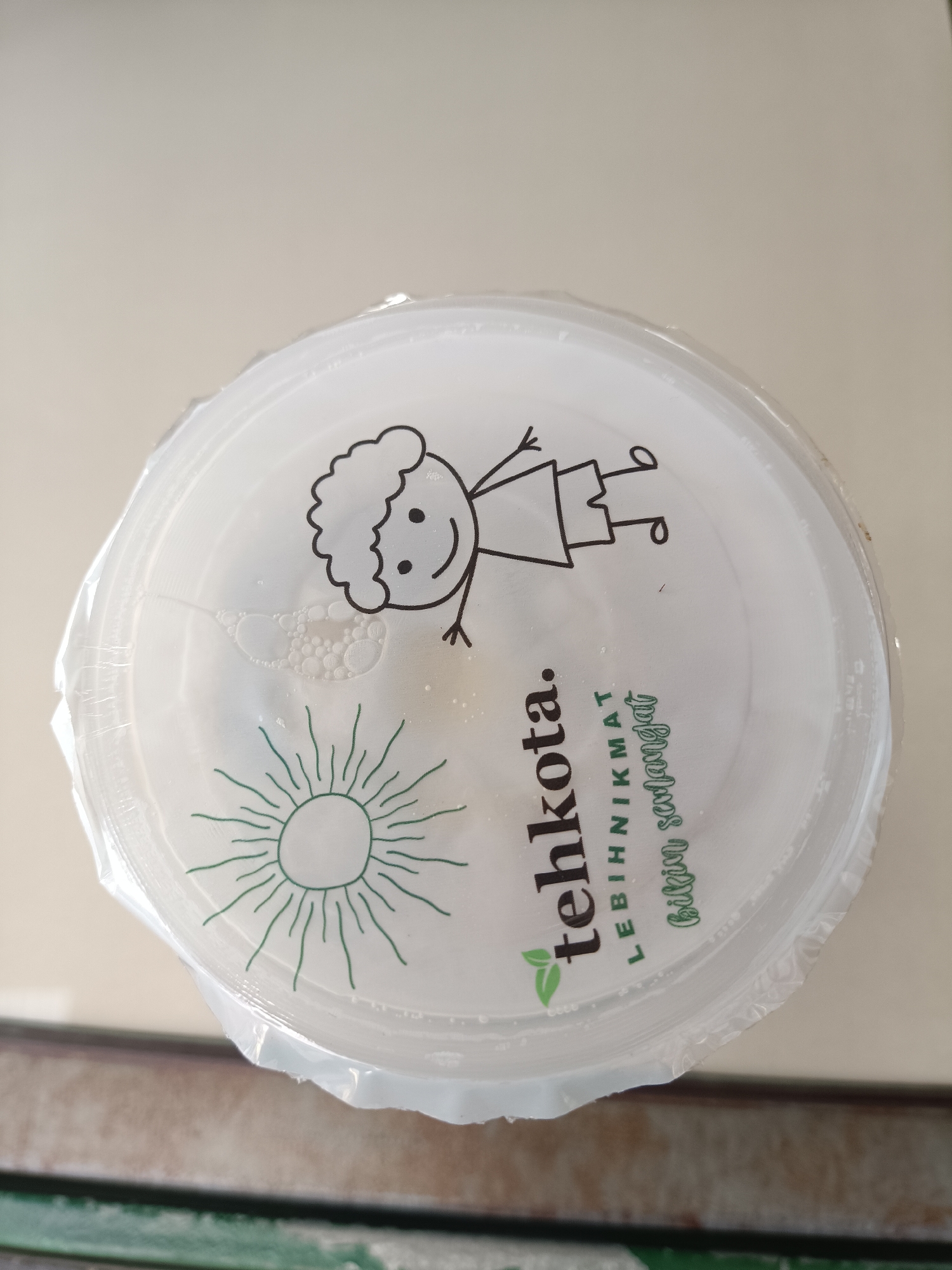 The image shows the top view of a clear plastic cup lid with a logo printed on it. The logo features a stylized drawing of a child with a bushy head, a smiling face, and stick figure arms and legs. To the left of the child drawing, there is a drawing of a sun with rays. In the center of the lid, there is text that reads "tehkota." Below "tehkota," there is smaller text that says "LEBIH NIKMAT" and "bikih sedapat". There are some small bubbles visible on the surface of the lid, suggesting it may have been recently filled with a beverage. The lid is sealed with clear plastic wrap. The background is a neutral, light-colored surface, possibly a table. There is no visible indication of the location being Jember, Indonesia.