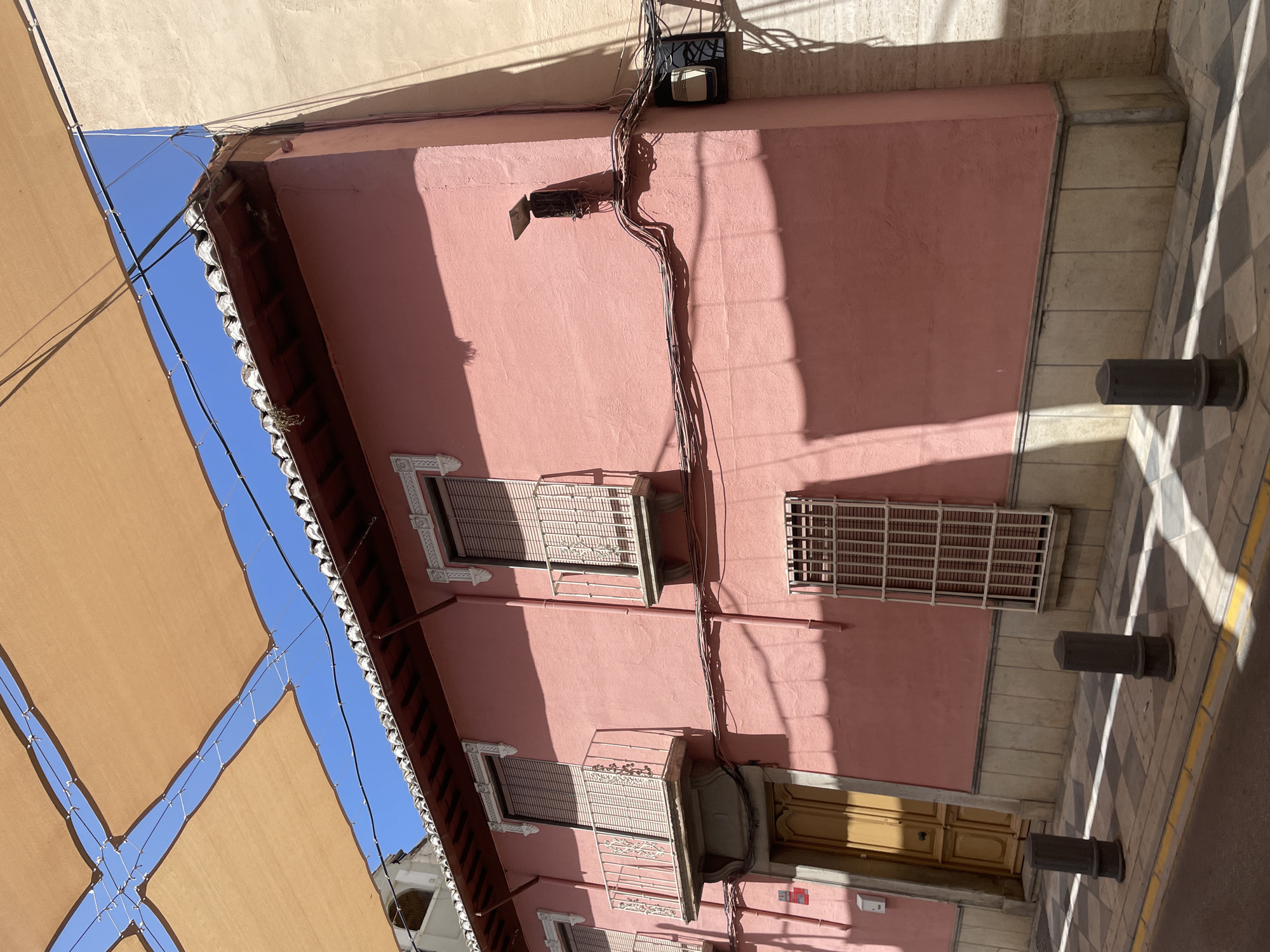 The image captures a street view in Atarfe, Spain, focusing on the facade of a pink building. The building has a red-tiled roof and white decorative elements around its windows and balconies. Two balconies with white railings are visible on the upper floor, and a barred window is on the lower floor. A light-colored wooden door is present on the ground level. The building's exterior is adorned with various electrical conduits and a light fixture mounted on the right side. Overhead, a tan sunshade is stretched across the scene, casting shadows on the building and the checkered pavement below. Several dark cylindrical bollards are placed along the sidewalk. The sky is visible between the panels of the sunshade.