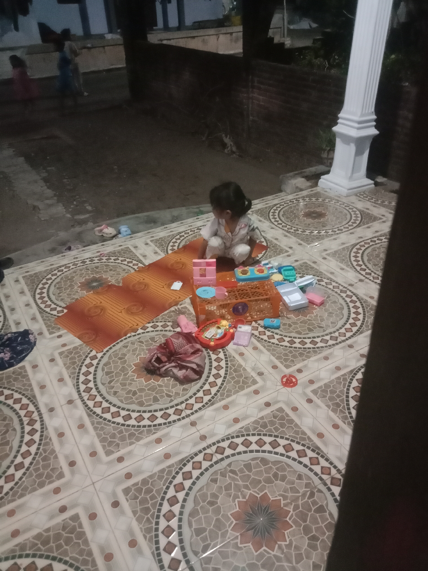 In Kedungsari, Indonesia, a young child is seated on a patterned tile floor, playing with various colorful toys. The toys include plastic building blocks and what appear to be miniature kitchen items, suggesting a play kitchen set. A patterned orange mat lies on the floor nearby. In the background, out of focus, several figures, possibly adults and other children, are visible, standing on what seems to be a dirt or gravel surface. A large white pillar is to the right of the scene, and a brick wall is visible further in the background. The lighting suggests it is nighttime or dusk.