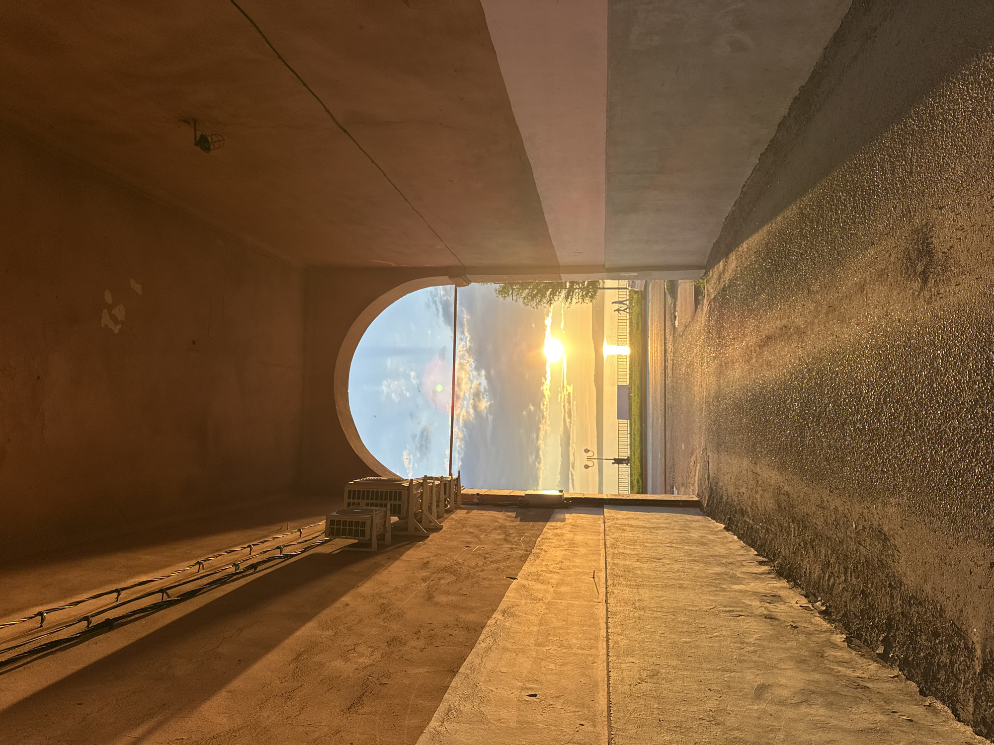 The image presents a striking view of a sunset through an arched underpass or tunnel, looking out towards a large body of water, likely the Votkinsk Reservoir in Votkinsk, Russia. The perspective is from inside the passage, which has concrete walls. The left wall, painted a warm reddish-brown, features an overhead light fixture and several parallel electrical cables running along it, casting prominent shadows. Below the cables, multiple external air conditioning units are mounted. The right wall and the floor, possibly asphalt or textured concrete, are bathed in the warm light from the setting sun.
Through the large, rounded archway, the sun is visible as a bright, intense golden orb, low on the horizon. It creates a long, shimmering path of light across the expansive water. The sky transitions from a clear blue with scattered white clouds on the left to vibrant oranges and yellows around the sun. Along the distant waterfront, a strip of land or road is visible, bordered by a railing or fence. Silhouetted trees stand to the left of the exit, and a decorative lamppost with two lights is present on the right. A faint human figure appears to be standing near the lamppost, observing the serene scene. The strong directional light and extended shadows clearly indicate it is late afternoon or early evening during sunset. No visible text is present.