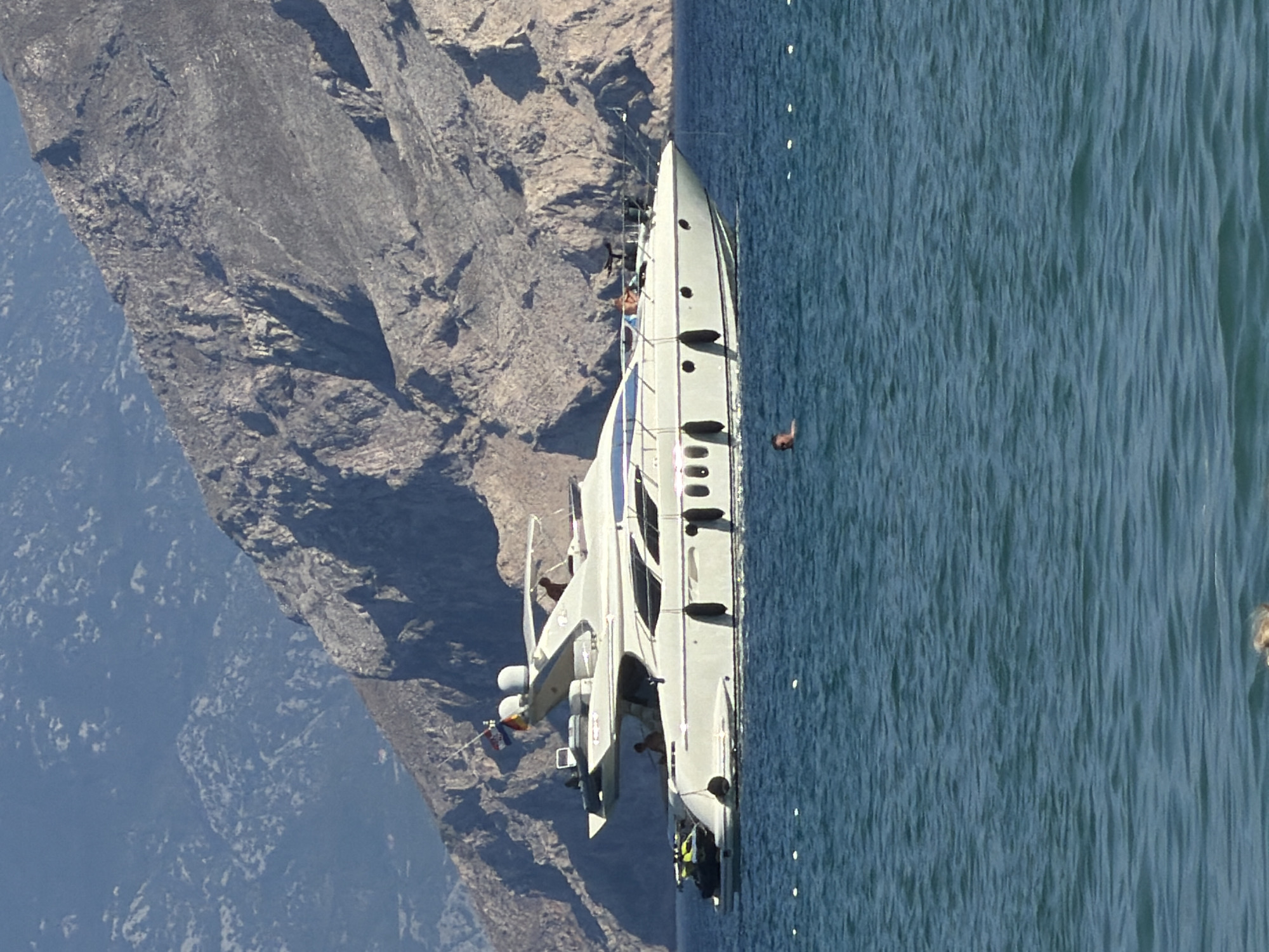 The image displays a modern, white yacht moving through calm, blue water alongside a steep, rocky mountainside. The yacht has several levels and appears to be a luxury vessel, with multiple windows visible along its side. There are a few people on the deck of the yacht, some of whom are wearing brightly colored clothing, suggesting a leisurely activity. One person is leaning over the railing. Another person is in the water near the yacht, with their arm raised as if waving or calling out. Small white buoys are visible in the water, arranged in a line. The sun appears to be high in the sky, casting a bright light on the scene, indicating it is daytime. The rocky terrain of the mountainside is rugged and barren, with some sparse vegetation visible on the lower slopes. The water is relatively still, with only small ripples. The overall impression is one of a pleasant day out on the water in a scenic coastal location. The image was taken in Općina Baška, Croatia.