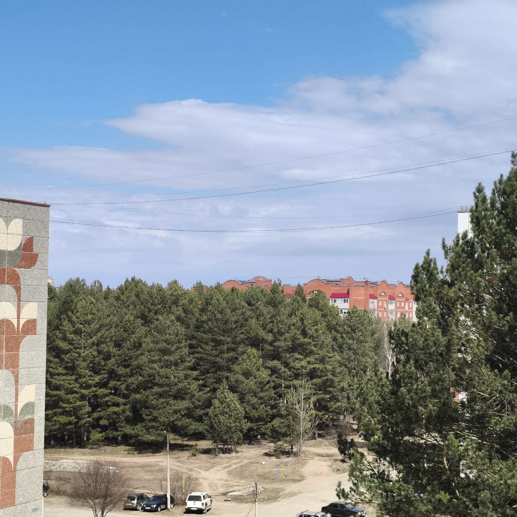 This media file presents an outdoor, elevated view of a residential area bordering a dense pine forest in Chaikovsky, Russia, captured during daylight hours on a clear, sunny day.
The scene is dominated by a vast forest of coniferous trees, filling the midground. Beyond the treeline, a cluster of multi-story buildings with distinctive red roofs is visible, suggesting a residential neighborhood. These buildings feature numerous windows and some have arched architectural details on their roofs. A lighter-colored building is partially obscured by trees on the far right.
In the immediate foreground, to the left, a section of a building with a unique geometric, mosaic-like pattern in shades of grey, brown, and cream occupies the frame. Below the forest, an unpaved area of dirt and sparse vegetation serves as a ground-level space where several cars are parked, including a dark SUV/van, a silver car, a white SUV, and a dark sedan. A street lamp and a small metal structure are also present in this area.
The sky above is bright blue with scattered white, cumulus clouds. Multiple utility lines traverse the upper portion of the image. No people or specific activities are visible, conveying a sense of calm and everyday life. No legible text is apparent in the image.