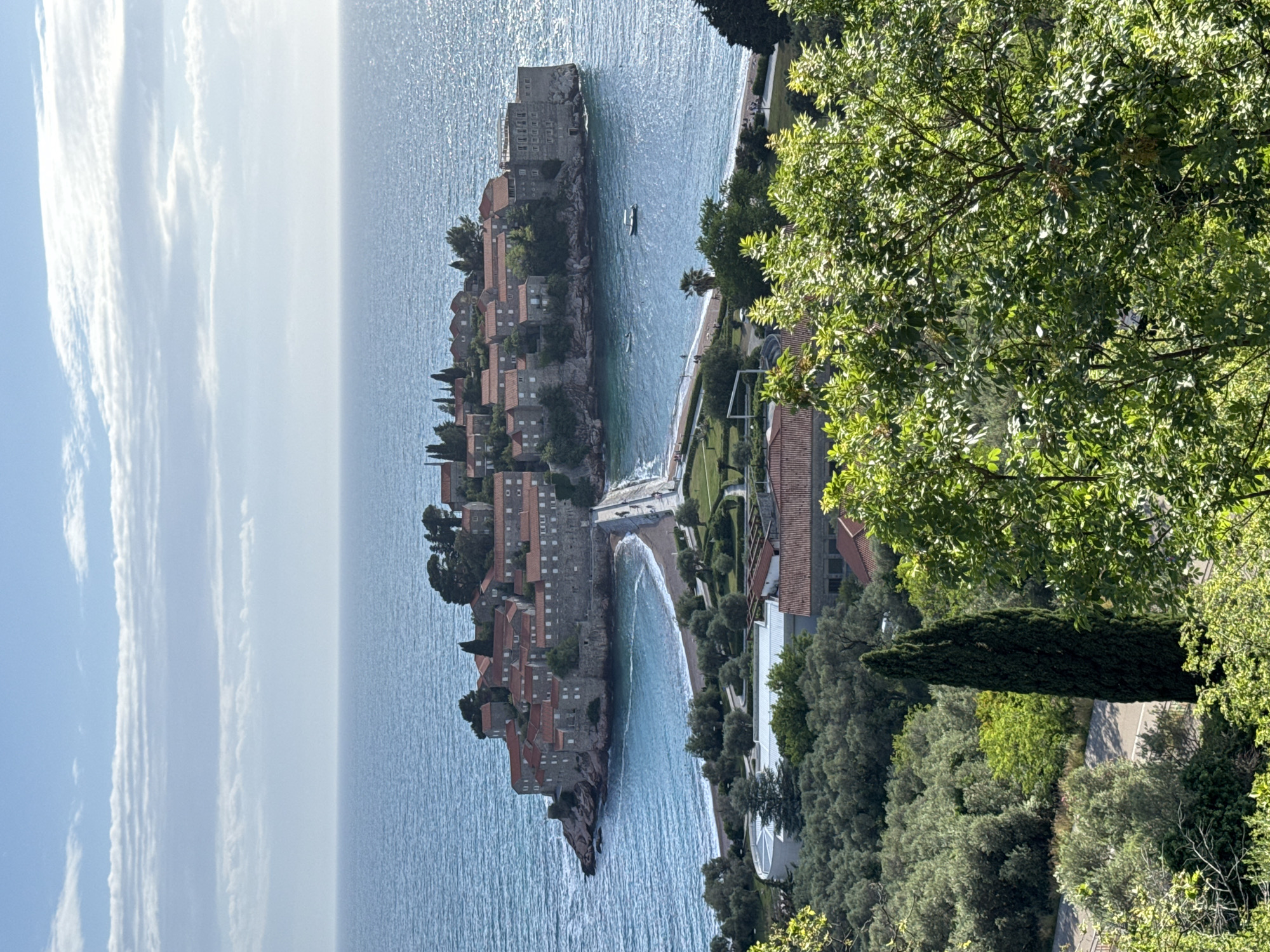 The image shows an aerial view of Sveti Stefan, Montenegro. The scene is dominated by the island of Sveti Stefan, a historic fortified settlement connected to the mainland by a narrow isthmus. The island is densely packed with stone buildings featuring terracotta-tiled roofs. The isthmus connects the island to a sandy beach. The sea surrounding the island is a bright, reflective blue. On the mainland, lush green vegetation covers the landscape. A paved road snakes through the trees. A modern building with an orange-tiled roof is also visible on the mainland. The sky is partially cloudy.