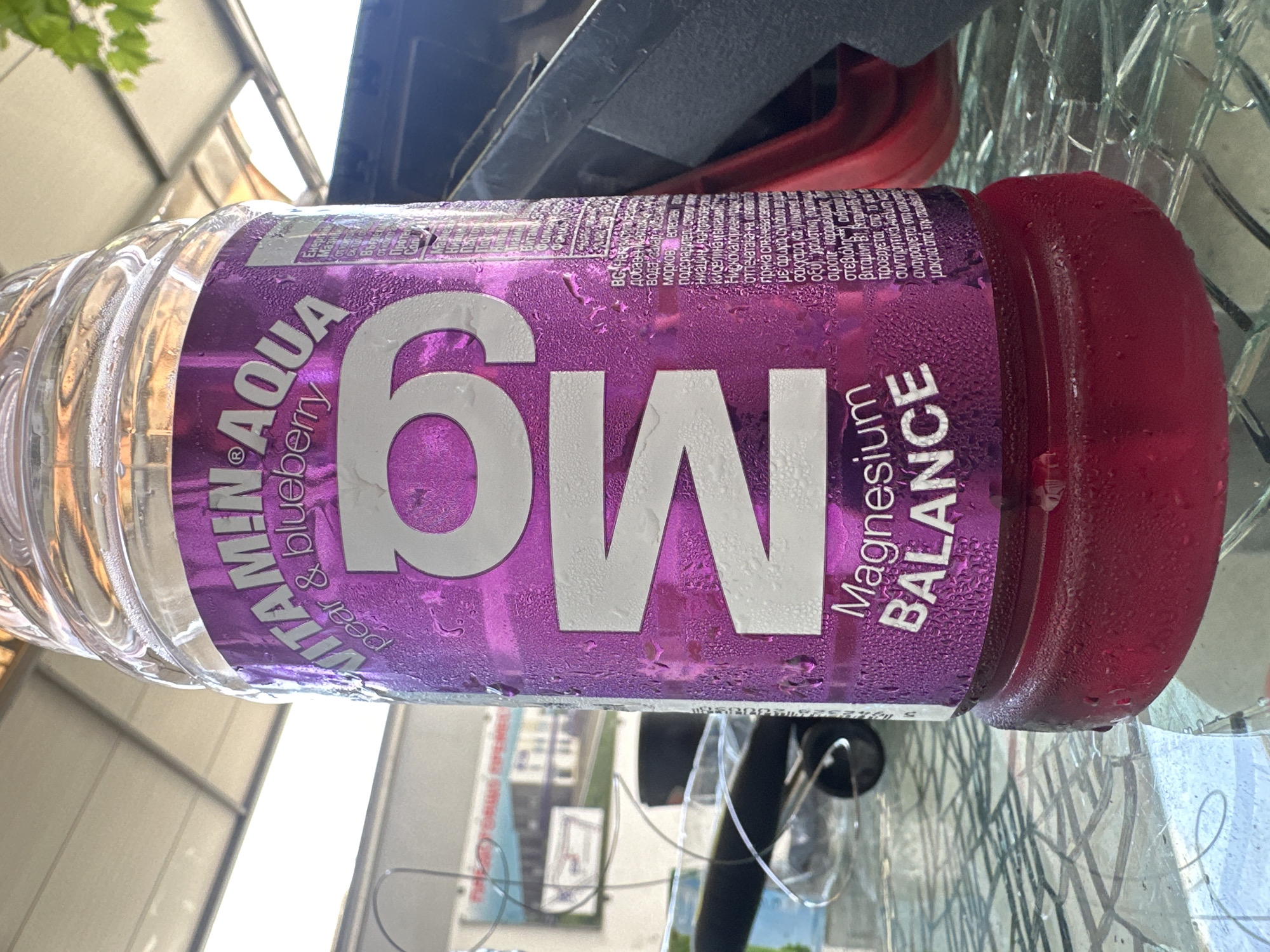 A close-up, slightly low-angle shot shows a clear plastic bottle filled with a reddish liquid. The bottle has a prominent purple label with "VITAMIN AQUA" and "pear & blueberry" printed in silver and white lettering. A large, stylized "M9" is also printed in white on the label. Below this, the text "Magnesium BALANCE" is visible in white lettering. The bottle is covered in condensation. The bottle rests on a glass surface with a geometric pattern, possibly a table. In the background, a metal awning or roof structure is visible, along with some green foliage. To the right of the bottle, a dark, possibly plastic object is partially visible. The overall scene appears to be outdoors.