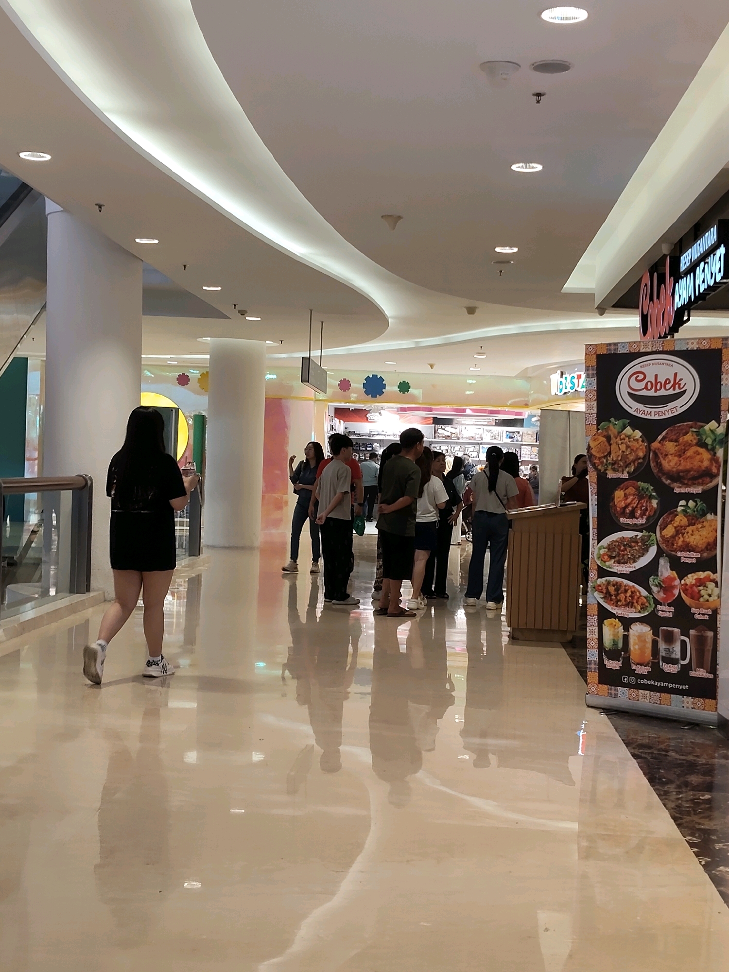 The image captures a scene inside a modern shopping mall in the City of Medan, Indonesia. The mall features a curved, white ceiling with integrated lighting and subtle cove lighting. Large white cylindrical pillars punctuate the spacious walkway, which has a highly reflective beige floor.
In the foreground, a woman with long, dark hair walks away from the camera, wearing a black t-shirt and shorts, and white sneakers. Her reflection is clearly visible on the glossy floor.
Further into the mall, a group of people are gathered, appearing to be interacting or waiting. Among them are individuals in casual attire, including a woman in a denim shirt and jeans, a man in a grey t-shirt and dark pants, and others standing near a wooden counter.
To the right of the frame, a large vertical banner displays images of food items and text in Indonesian, advertising a restaurant or food stall named "Cobek Ayam Penyet." The banner features pictures of various dishes, including fried chicken, stir-fried items, and drinks, along with the restaurant's logo and social media handle. The overall ambiance suggests a public indoor space, likely a food court or a common area within the mall.
