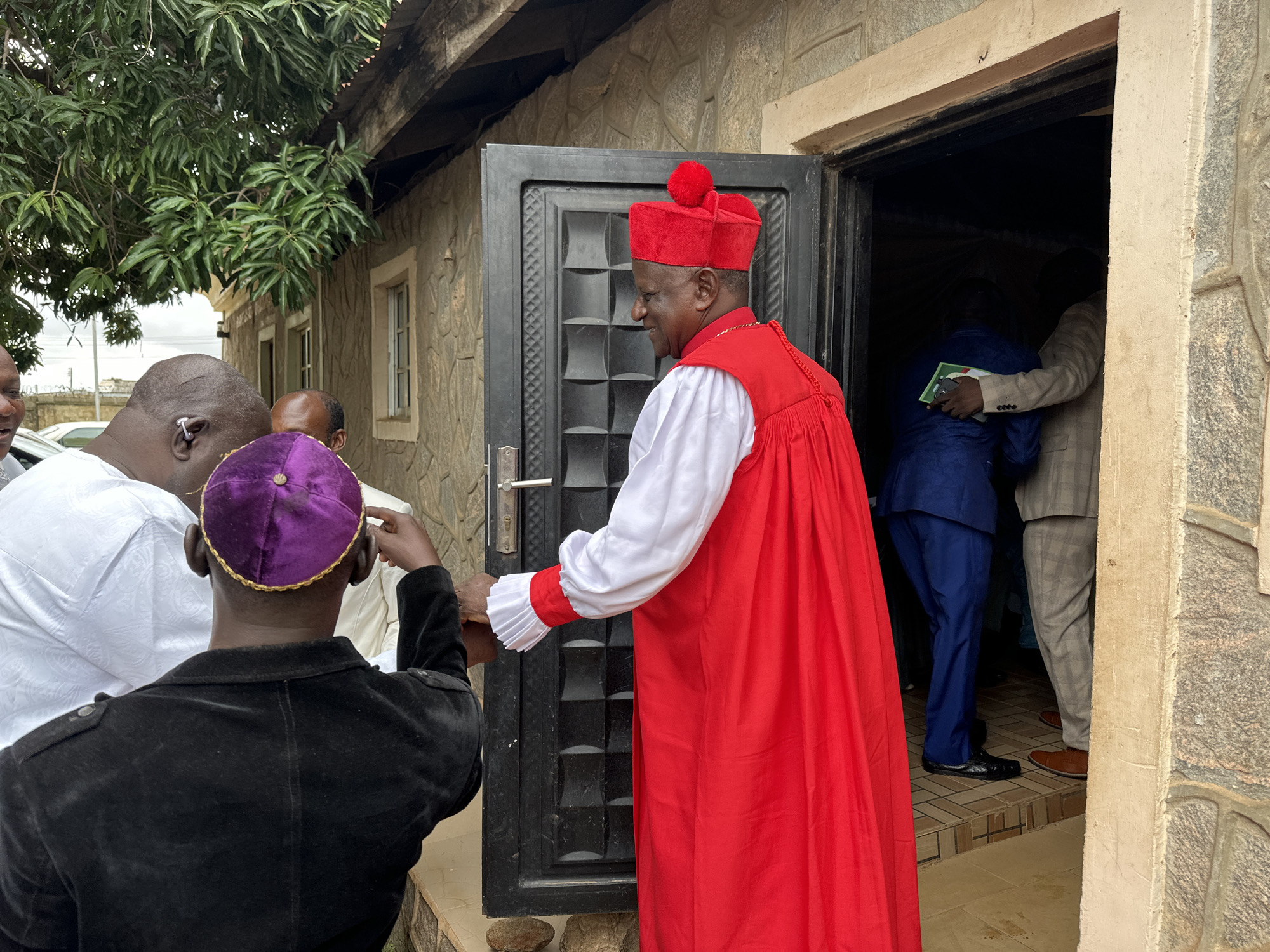 In Barden Yana, Nigeria, a group of men gathers outside a building with a stone facade. A man in a red ceremonial robe and matching hat, adorned with a red pom-pom, stands in a doorway, facing right and smiling. He is interacting with a man in a black jacket and purple cap. To the left of the doorway, a man in a white garment is partially visible. Inside the doorway, two other men are seen; one in a blue suit and another in a plaid suit. The building has a dark metal door with a textured surface. Trees with dense green foliage are visible overhead, and the ground appears to be paved. The scene suggests a formal gathering or religious event.