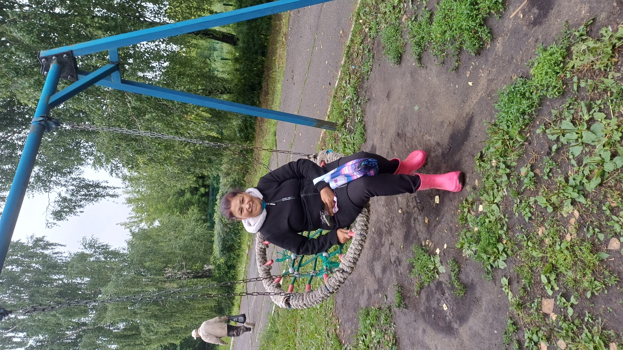 In Podgornoye, Russia, a middle-aged woman of indeterminate ethnicity is seated on a circular rope swing. She is wearing a black zip-up hoodie with a light-colored hood, black pants, and bright pink rubber boots. She holds a smartphone and a patterned fabric bag in her lap. The swing is suspended from a blue metal frame. In the background, a paved path runs alongside a grassy area, and trees line the perimeter. Another person, wearing a light-colored jacket and dark pants, is walking away in the distance on the path. The ground around the swing is a mixture of dirt and sparse green vegetation.