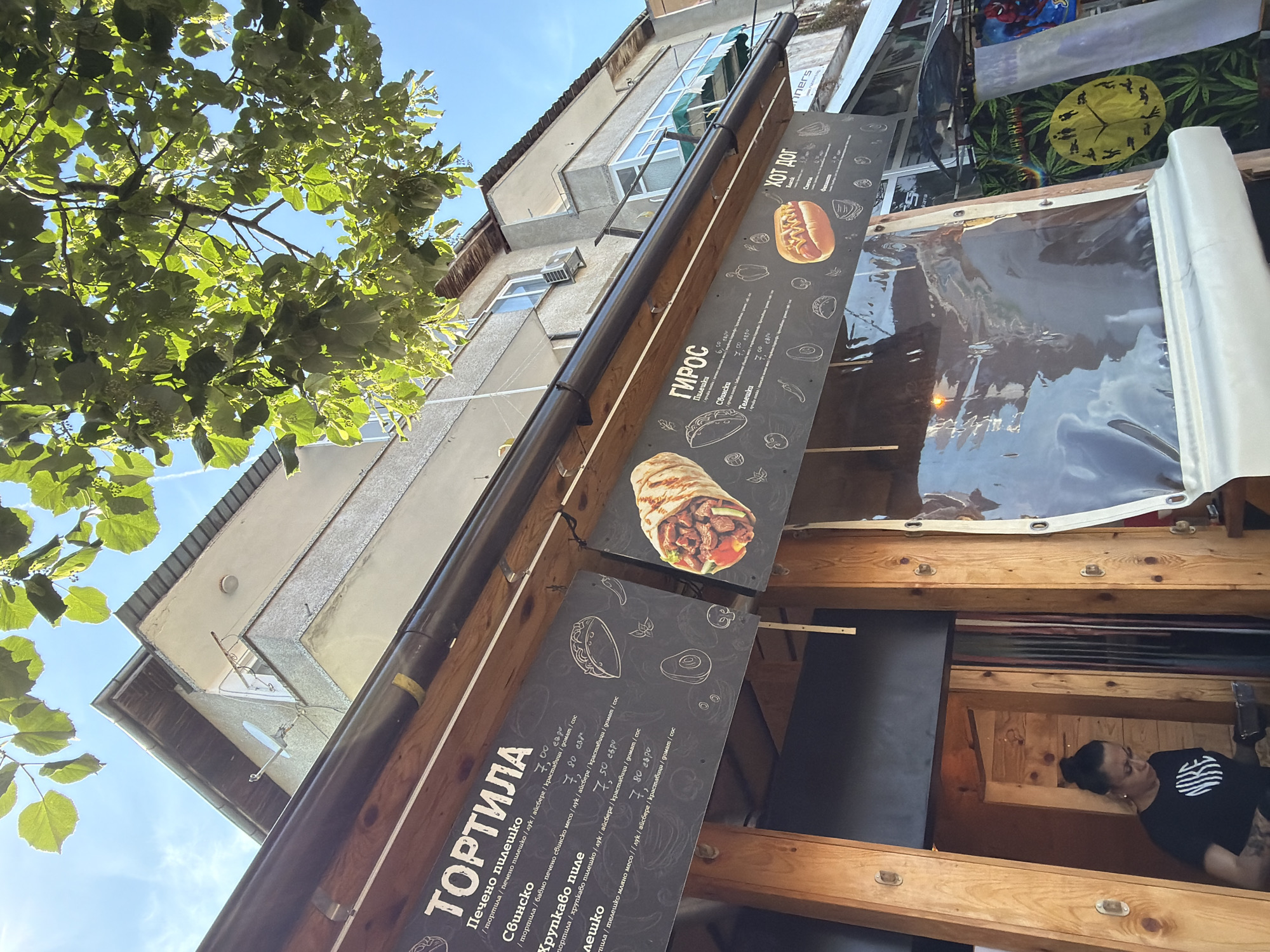 The image shows a street-level view of a food stand or cafe, likely in the city of Tsarevo, Bulgaria, given the Bulgarian text on the menu boards. Sunlight streams down from a clear blue sky, illuminating the scene and casting shadows. Lush green leaves from an overhanging tree partially obscure the upper part of the image.
Dominating the foreground are two large, black menu boards suspended from a wooden awning. The left board is titled "ТОРТИЛА" (Tortilla) and lists various tortilla options with their ingredients and prices in Bulgarian leva. Examples include "Печено пилешко" (Roast chicken) and "Свинско" (Pork). To the right of the "Tortilla" menu, a detailed illustration of a wrapped tortilla is visible.
The right menu board is titled "ГИРОС" (Gyros) and also displays menu items and prices in Bulgarian. Below the "Gyros" menu, there is a picture of a hot dog with mustard, labeled "ХОТ ДОГ" (Hot Dog). Further to the right, a partial view of another sign with "ers" visible at the bottom suggests it might be a business name.
In the lower left quadrant of the image, a woman wearing a black t-shirt with a white "NIKE" logo is visible inside the food stand. She appears to be working, possibly preparing food, and has her hair tied up in a bun. Her arms are adorned with tattoos.
The background features a multi-story building with windows and balconies, typical of urban architecture. A satellite dish is mounted on the side of the building, and an air conditioning unit is visible on one of the balconies. The overall impression is that of a casual, open-air food establishment operating on a sunny day.