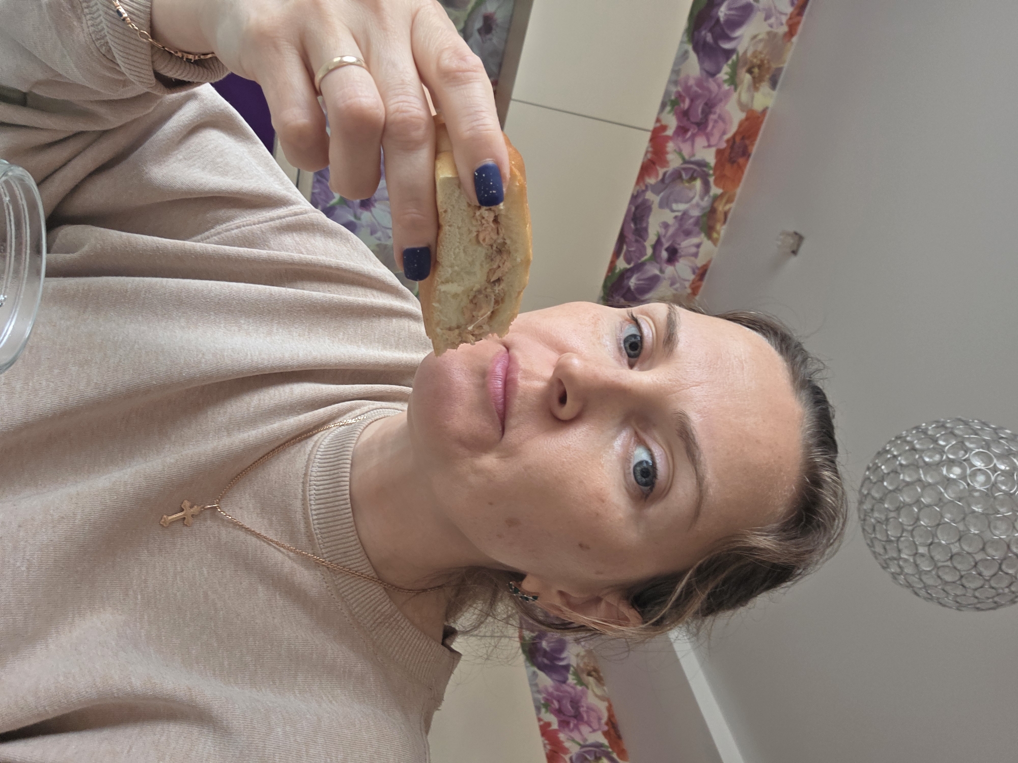 A low-angle, rotated photograph captures a person indoors, holding a piece of food towards their mouth. The subject, likely female, has fair skin, light blue eyes, and light brown hair. She is wearing a light beige or tan long-sleeved sweatshirt. A thin gold-colored chain with a small cross pendant is visible around her neck, and a gold ring is on her left ring finger. Her fingernails are painted dark blue with subtle glitter.
She is holding a small, light-colored bun or pastry that appears to be filled with a savory mixture, possibly tuna or finely ground meat, positioning it just below her lips. Her eyes are wide and focused directly on the camera, and her expression is neutral.
The background features an interior space. On the left side and behind the subject, a wall is decorated with floral wallpaper displaying patterns of purple, orange, and green flowers. The upper right portion of the image shows a plain, light-colored wall or ceiling. A spherical light fixture, constructed from clear, bubble-like elements, hangs prominently on the right side of the frame. A clear glass object, possibly a bowl, is partially visible in the bottom left corner. The image depicts a casual moment, possibly during a snack or meal in Chaikovsky, Russia.
