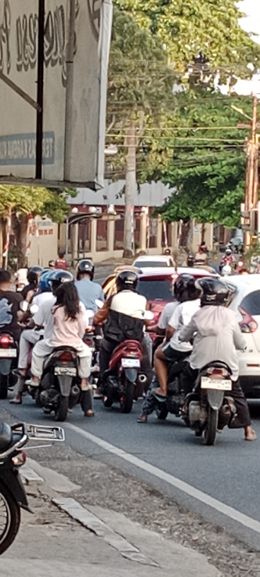 The image depicts a street scene in Tanjung Pinang, Indonesia, likely during daylight hours given the lighting and shadows. Multiple motorcycles are present, forming a traffic jam on a paved road. Most of the individuals are wearing helmets, with some passengers visible. The motorcycles are of various models and colors, with some carrying one or two people. The background shows buildings with reddish-brown accents, trees with lush green foliage, and utility poles with overhead wires. The road appears to have a white dividing line and a worn curb on the side, with some debris and cracked pavement. The overall impression is of a busy urban environment during a period of congestion.