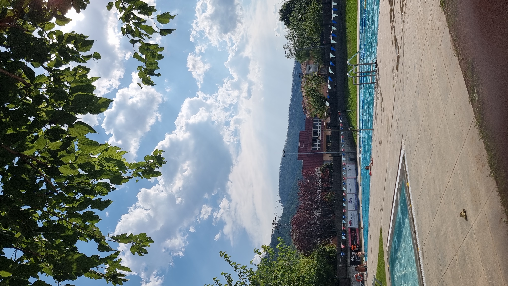 The image depicts an outdoor swimming pool complex under a partly cloudy blue sky, framed by green foliage from overhead trees. The complex features multiple swimming pools, with one in the foreground and a larger one in the mid-ground. The larger pool is lined with blue and white triangular flags and has several people swimming or relaxing. In the background, there is a red building with white window frames, surrounded by trees and hills. The paving around the pools is light-colored concrete. The scene appears to be located in Sant Joan Les Fonts, Spain, given the presence of a sign with text that likely corresponds to local establishments.