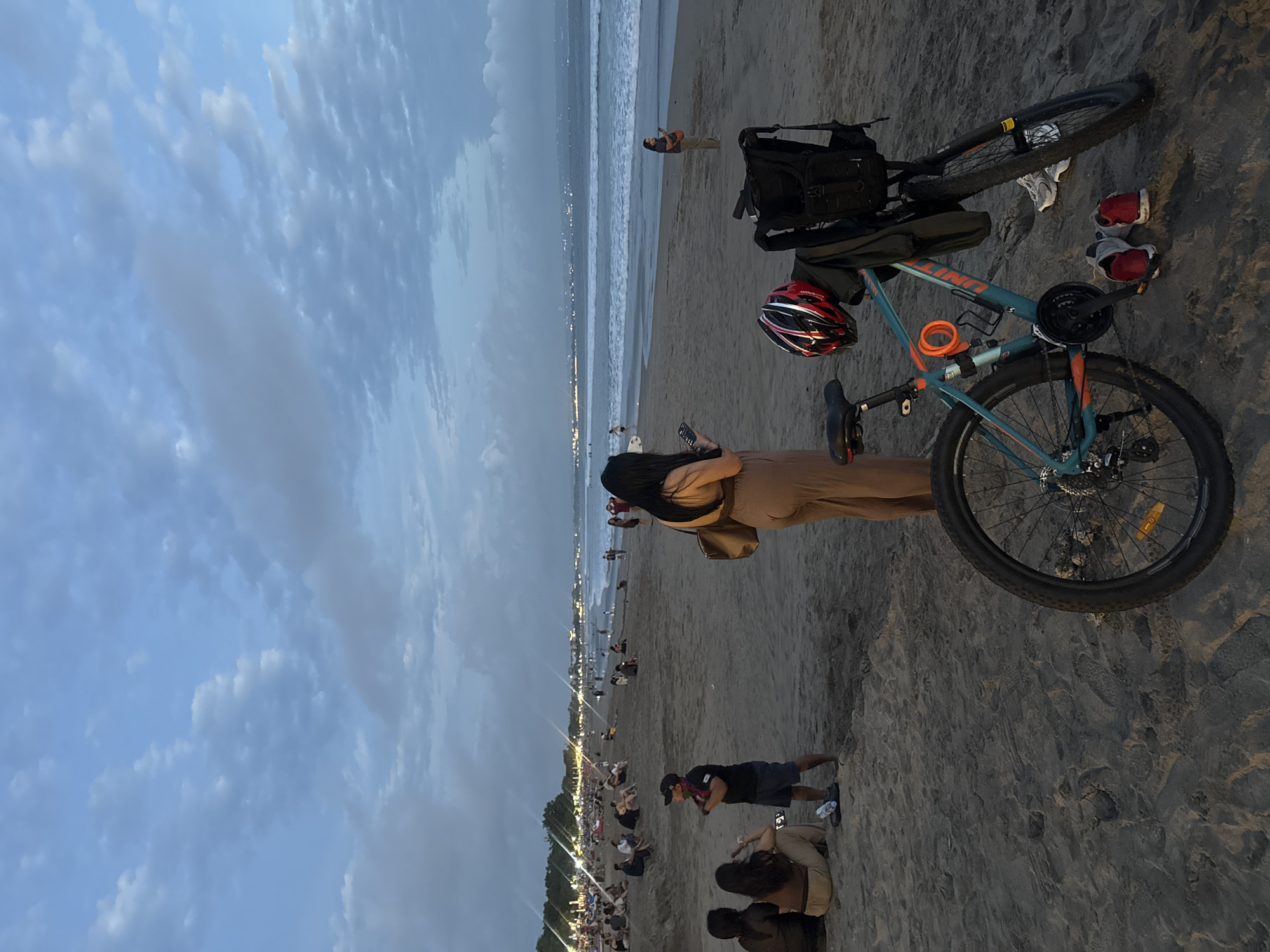 In Kuta, Indonesia, at dusk, a beach scene unfolds with a teal bicycle parked on the sand in the foreground. The bicycle is equipped with a red and black helmet attached to the handlebars and a black backpack slung over the rear. A pair of red and white sneakers rests on the sand beside the bicycle.
In the middle ground, a woman wearing a tan jumpsuit stands with her back to the camera, looking at her phone. Further down the beach, people are scattered across the sand, some sitting and others walking. The ocean stretches to the horizon, with gentle waves breaking on the shore. In the background, the lights of the city are visible, and the sky is a soft blue with scattered clouds, suggesting the transition from day to night. The overall atmosphere is one of relaxed evening leisure.