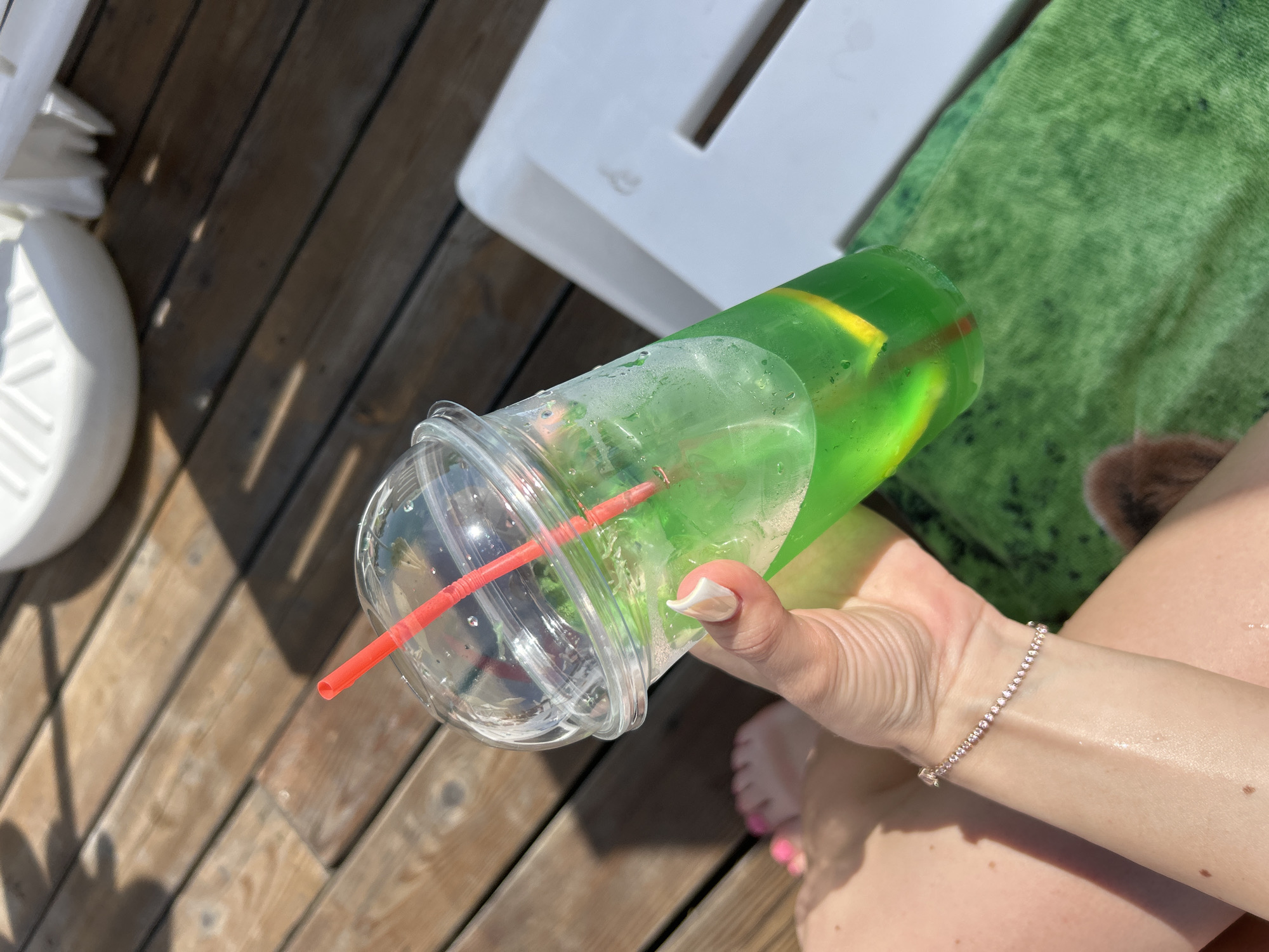 A first-person view shows a person's right hand holding a cold green beverage in a clear plastic cup with a domed lid and red straw, while reclining outdoors in Unknown, Unknown. The drink contains ice and yellow citrus slices. The person's right hand has a white manicured thumbnail, and a delicate, sparkling bracelet adorns the right wrist. Partially visible in the upper left, the person's left foot has pink painted toenails. The setting features light brown wooden decking under direct sunlight, casting shadows. A white plastic outdoor chair or lounger armrest is visible on the right, and a textured green towel is draped over the person's lap. In the blurred background, a white, possibly ribbed, object is partially visible.
