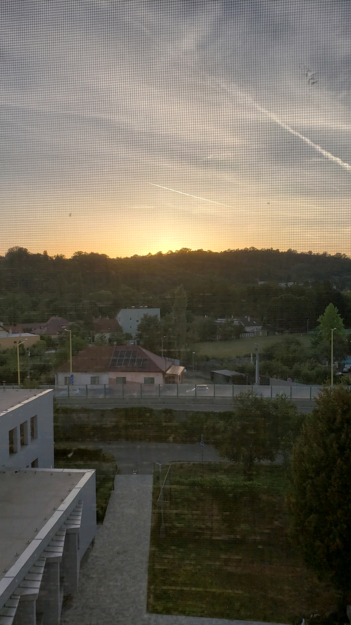 The image captures a sunset scene viewed through a screened window, likely from an elevated position, looking out over a suburban or semi-rural area. The sky is a gradient of pale yellow and orange near the horizon, transitioning to a soft blue and grey higher up. A distinct contrail from an aircraft is visible diagonally across the upper portion of the sky.
In the middle ground, a treeline forms a dark silhouette against the sunset, with a few houses and buildings nestled amongst the trees. One building in particular, a residential house with a reddish-brown roof, is notable for having solar panels installed. Another lighter-colored building, possibly an apartment block or office, is visible to the left of this house. A road runs horizontally across the middle of the image, with a few cars visible, suggesting moderate traffic. A fence runs along the side of the road.
In the foreground, the edge of a modern building is visible on the left, with a flat roof and geometric architectural elements. Adjacent to this building, a paved pathway leads downwards towards a grassy area. The overall impression is one of a tranquil, late afternoon or early evening view, potentially from a residential building or a commercial establishment overlooking the outskirts of a town.
Based on the presence of the distinct architectural styles of the buildings, particularly the modern building in the foreground and the residential structures with solar panels, combined with the landscape, this scene is likely located in the city of Prešov, Slovakia. The combination of urban development and the surrounding natural landscape is characteristic of the city's setting.
