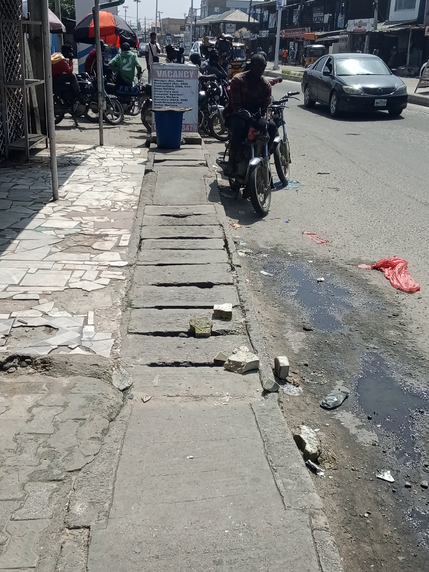 In Ado, Nigeria, a street scene is depicted on a bright, sunny day. The foreground features a cracked and uneven concrete sidewalk with scattered debris and broken tiles. A concrete drainage channel runs alongside the sidewalk, also showing signs of wear and tear.
To the right of the sidewalk, the street is visible with a dark, murky puddle of water and some refuse, including a crumpled pink plastic bag and other litter. Further down the street, several motorcycles are parked. One motorcycle in the mid-ground has a man with a red and black checkered shirt sitting on it, facing away from the camera. Behind him, a white and blue sign advertising "VACANCY" is displayed, listing various job positions.
Across the street, a dark-colored car drives by. Buildings line the opposite side of the street, with awnings and shops visible. People are seen walking and standing on the sidewalks of the buildings across the street. The overall impression is one of a bustling urban environment with signs of aging infrastructure and casual littering.