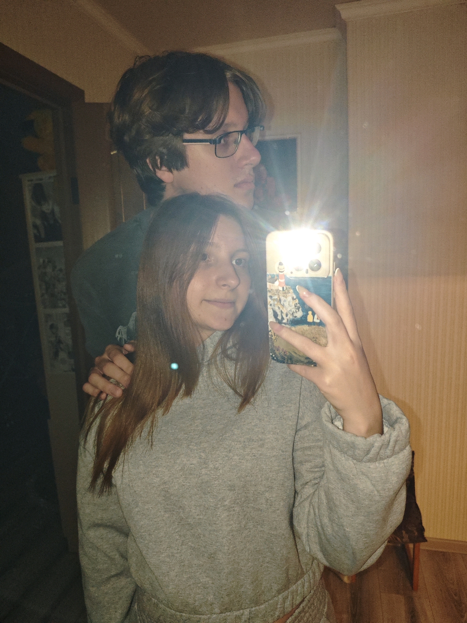 In a close-up, indoor selfie, a young woman in a grey hoodie holds a smartphone with a scenic phone case, its flash illuminating her face. Her hair is long and brown, and she has a gentle smile. Behind her, a young man with brown hair and glasses is partially visible, with his hand resting on her shoulder. The background appears to be a room with patterned wallpaper and a wooden floor. The overall lighting is warm, likely from artificial sources. No specific Vawkavysk landmarks or features are discernable.