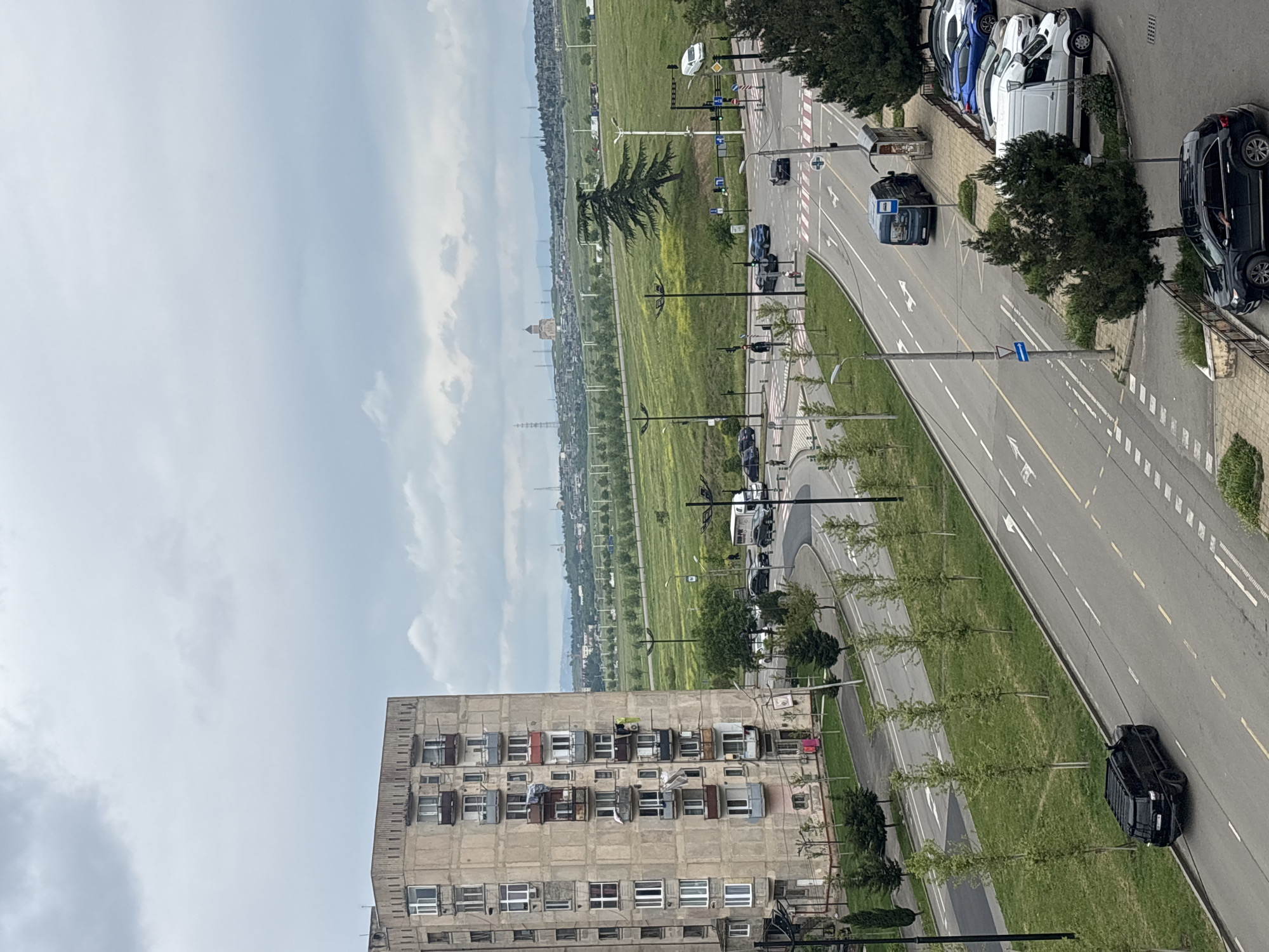 The image shows an aerial view of a street in Tbilisi, Georgia, likely taken from a high vantage point within an apartment building. The perspective is slightly tilted.
The foreground features a large, weathered concrete apartment building with multiple rows of windows, some with laundry hanging out. A street runs alongside this building, marked with painted lines and arrows indicating traffic direction. Several cars are parked along the street, and a few are in motion.
Beyond the road, a wide expanse of green landscape stretches into the distance, punctuated by scattered trees, light posts, and possibly pedestrian walkways. In the background, there's a silhouette of mountains or distant hills under a cloudy sky. A distinct structure resembling a tower or dome is visible on the horizon.