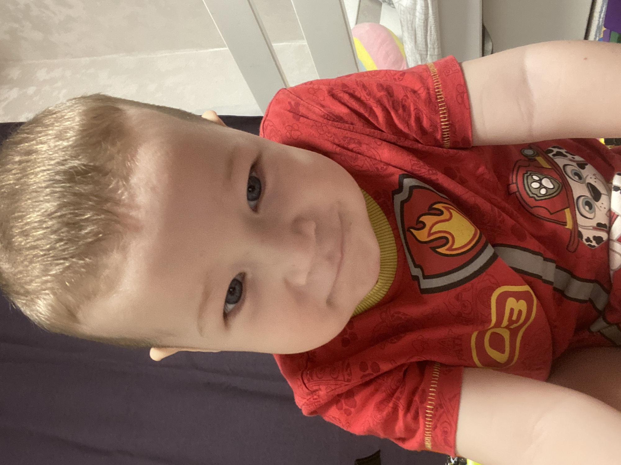 A young child with fair skin and blond hair is lying down, possibly on a bed, wearing a red t-shirt with a "Paw Patrol" design. The background includes what appears to be a white crib or bed frame, along with some bedding or pillows. The location is not discernible from the image.