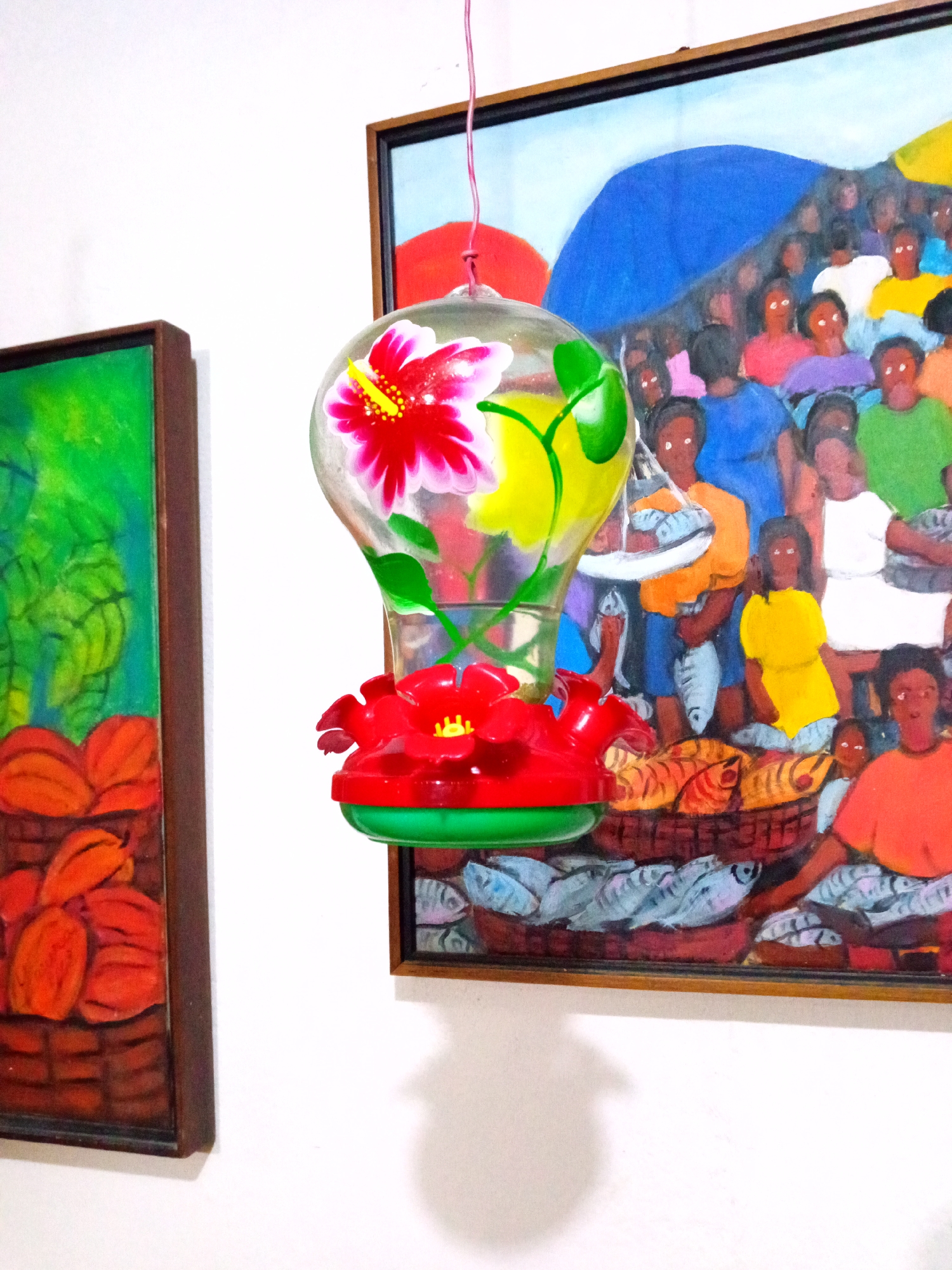 A hummingbird feeder hangs in front of two framed paintings. The feeder is clear with a red and green base and a red hibiscus flower design on the upper portion. The painting on the right depicts a market scene with people holding fish and fish in baskets. The painting on the left shows what appear to be cacao pods in a basket. The scene is indoors with a white wall in the background. Although the location is claimed to be El Limón, Venezuela, this cannot be determined from the imagery.