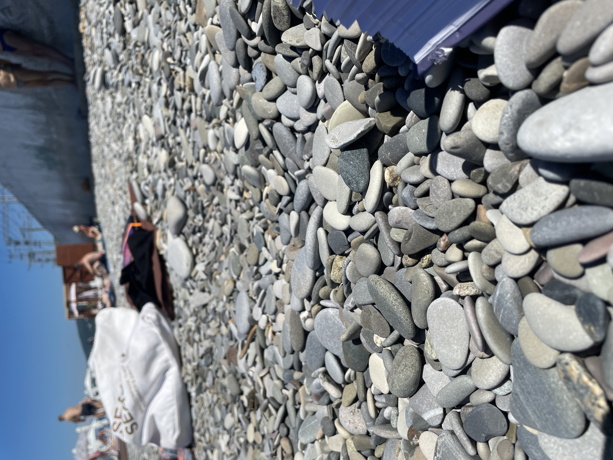 The image depicts a sun-drenched, rocky beach with a shallow depth of field. The foreground is dominated by a multitude of smooth, grey and white pebbles, some of which are partially obscured by a purple object in the bottom right corner. In the midground, a white cloth bag with gold lettering and a black item of clothing are scattered amongst the stones. Further in the background, several people can be seen relaxing on the beach, with some appearing to be swimming or wading in the water. The backdrop features a concrete wall, some wooden structures, and what appears to be a clear blue sky with a hint of industrial elements in the distance. The overall scene conveys a sense of a leisurely day at the seaside.