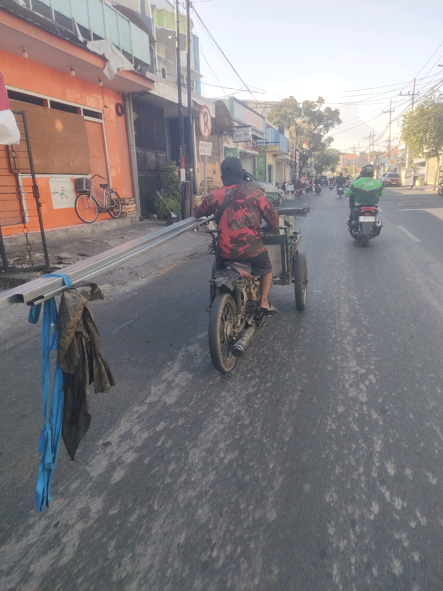 The image captures a street scene in Surabaya, Indonesia, likely during daytime given the bright sunlight and shadows. In the foreground, a modified motorcycle with a sidecar carries a person wearing a red and black camouflage shirt and dark shorts, riding on the right side of the road. The motorcycle is transporting a long, metallic pole, secured to the sidecar with blue straps and draped with a dark cloth. The road surface is asphalt, with visible patches of wear and tear.
Further down the road, other vehicles, including motorcycles, are present, suggesting moderate traffic. One motorcycle in the middle ground is ridden by a person wearing a green jacket, possibly a delivery rider, with a "Grab" logo visible. Buildings line the left side of the street, displaying a mix of architectural styles, including an orange-colored building with a bicycle parked in front of it and wooden boards covering part of a storefront. Power lines and utility poles are visible overhead. The overall impression is a typical urban environment in Indonesia, with a focus on the transport of goods via modified motorcycles.