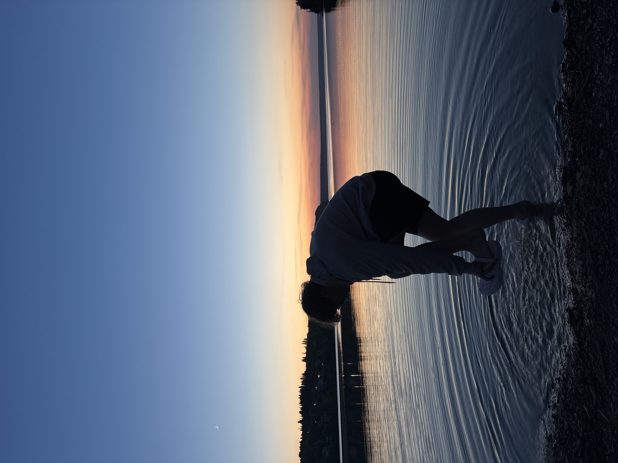 A person stands on a shoreline in what appears to be Gora (Khokhlovskoe S/P), Russia, based on the presence of a body of water and shoreline vegetation characteristic of the region. The person is wearing a hooded sweatshirt and shorts, with what appear to be shoes on their feet. They are leaning forward, partially submerged in the water. The body of water stretches into the distance, reflecting the sunset colors in the sky. A thin strip of land with trees or buildings lines the horizon. The sky transitions from a deep blue to orange and yellow near the horizon.