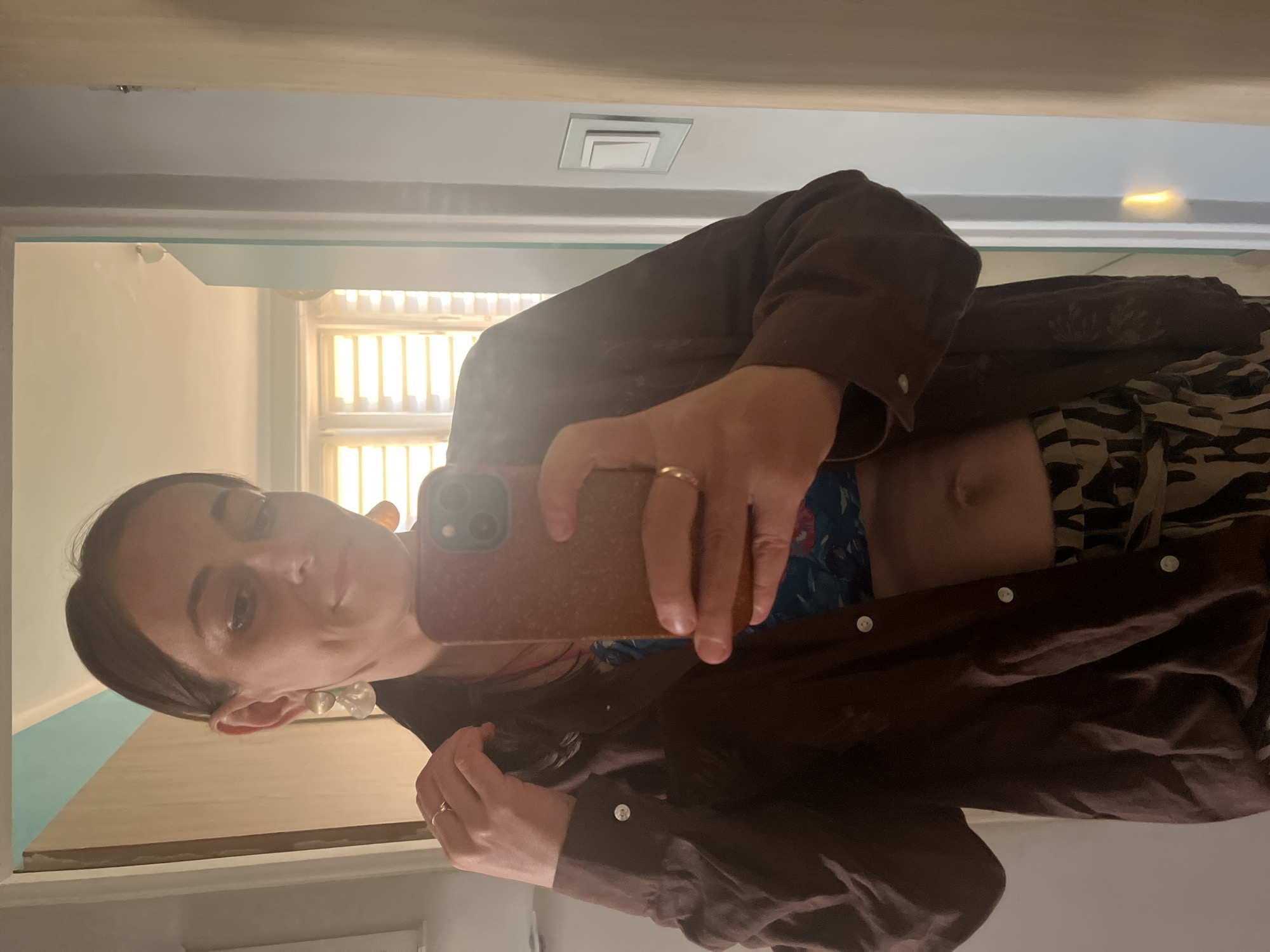 A woman is taking a selfie in a mirror. She is wearing a brown button-up shirt, a blue and floral patterned top, and a pair of patterned pants. She is holding a phone in her right hand and has her left hand on her hair. She is also wearing earrings and two rings. The background shows a room with a window and a light switch.