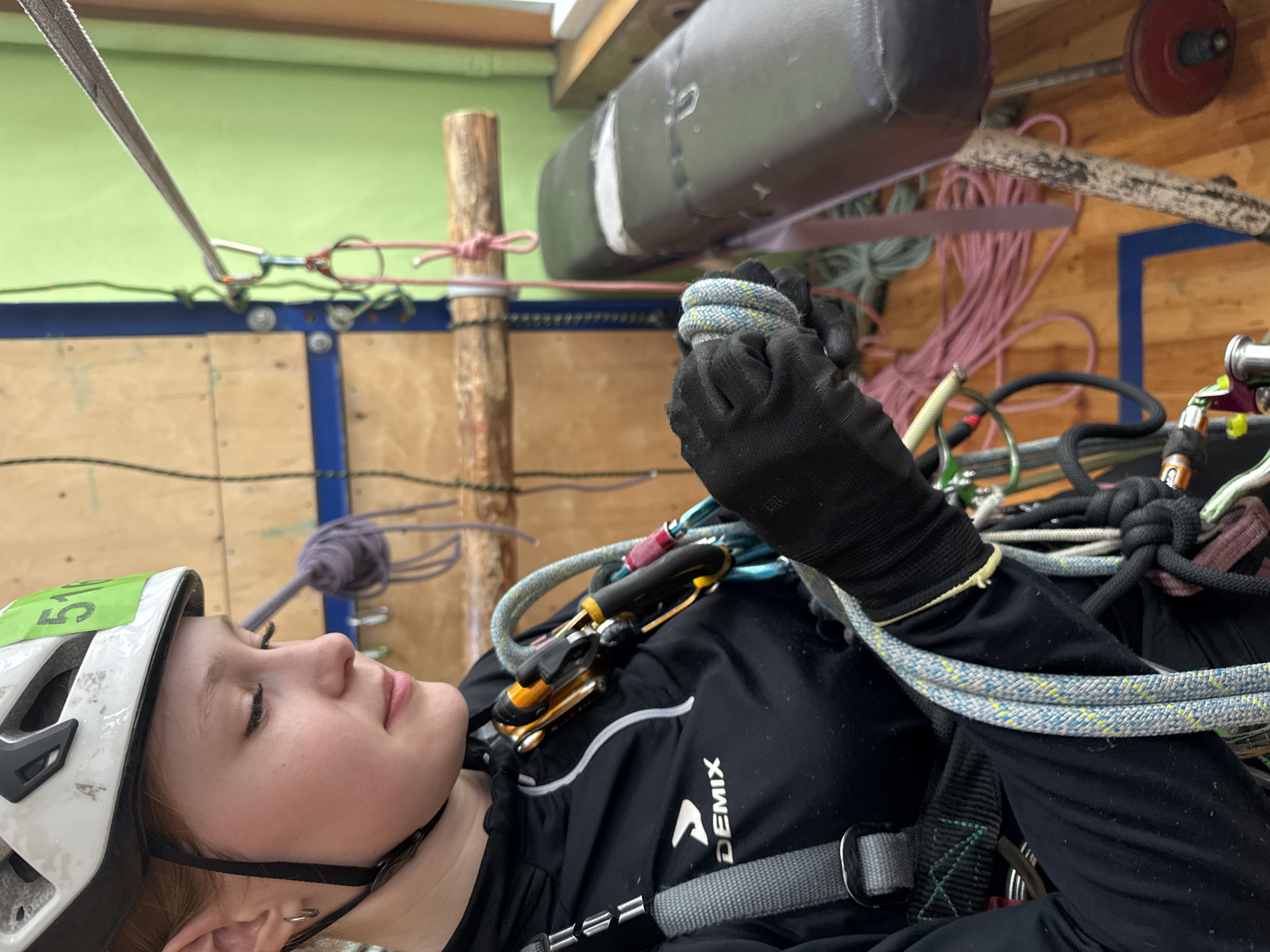 A person wearing a climbing helmet and a harness is shown in a seemingly indoor setting. The person is holding a coiled rope with gloved hands. Various other climbing ropes, carabiners, and related equipment are visible in the background. The location appears to be an indoor climbing facility. A logo of "DEMIX" is seen on the person's clothing. The specific location is Chaikovsky, Russia is not identifiable in this image.