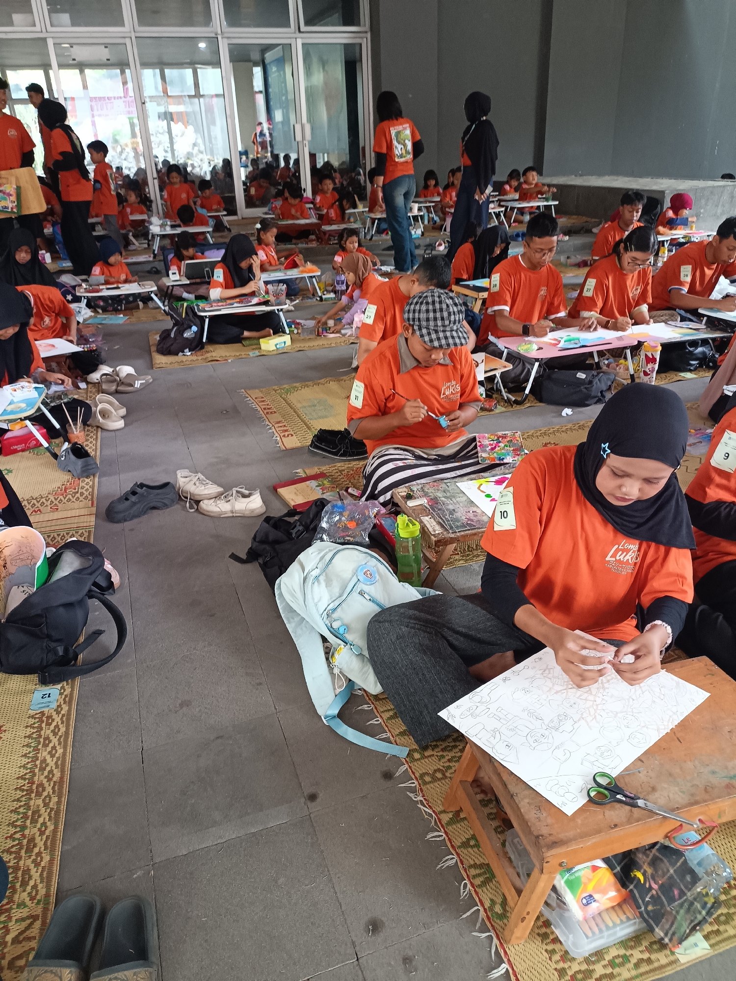 The image depicts an outdoor art competition or workshop for children and young adults in Hargobinangun, Indonesia. The scene is set on a paved ground area, likely a public space or courtyard, covered with woven mats. Numerous participants, predominantly in orange t-shirts, are seated on the ground, engaged in drawing and coloring activities at small, portable tables. The participants range in age and are wearing various attire, including hijabs for some female participants.
Objects present include drawing supplies such as colored pencils, markers, paint palettes, scissors, and sketchpads. Portable wooden tables and some backpacks are also visible. The background suggests an open-air venue with large glass-fronted buildings and possibly some overhead structures or awnings, indicating a shaded area. The lighting appears to be natural daylight, casting soft shadows. The overall atmosphere seems to be one of focused activity and community engagement.
