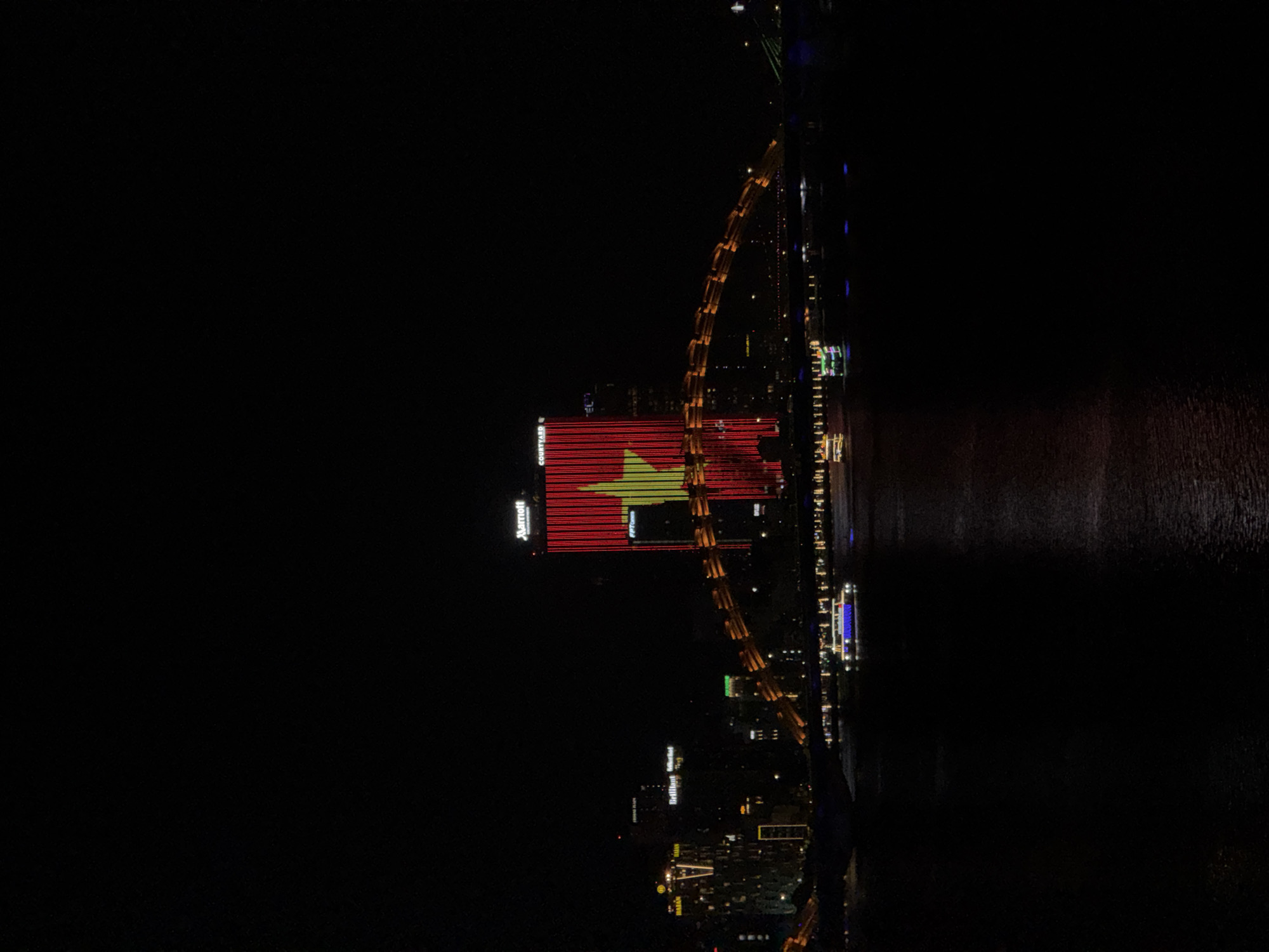 The image presents an illuminated urban nightscape, captured from an elevated perspective and oriented vertically. On the left side of the frame, a prominent tall building displays a large, brightly lit rendition of the Vietnamese flag – a yellow five-pointed star centered on a red background – across its facade. Vertically aligned text spelling "Marriott" is clearly visible in white letters above the flag display, and "COURTYARD" appears below it.
Adjacent to this building, a distinctive architectural structure, resembling a bridge, stretches vertically through the scene. This structure is brightly lit with warm yellow and orange lights, segmented into arch-like sections, strongly suggesting the Dragon Bridge, a famous landmark in Đà Nẵng.
To the right of the illuminated bridge, a dark body of water runs vertically along the entire right edge of the image. This water reflects the various city lights, indicating a calm surface, likely a river flowing through the city. Along the banks of this waterway and in the background, numerous other city buildings are visible, adorned with various artificial lights, creating a bustling urban panorama. Faint text on some of these other buildings includes "Brilliant Hotel," "NOVOTEL," and "A LA CARTE."
The scene captures a vibrant and well-lit city at night, emphasizing national pride through the prominent flag display and showcasing iconic architectural features. The setting is clearly a modern city, consistent with Đà Nẵng, Vietnam, at nighttime, with no discernible people or specific activities other than the general urban glow.