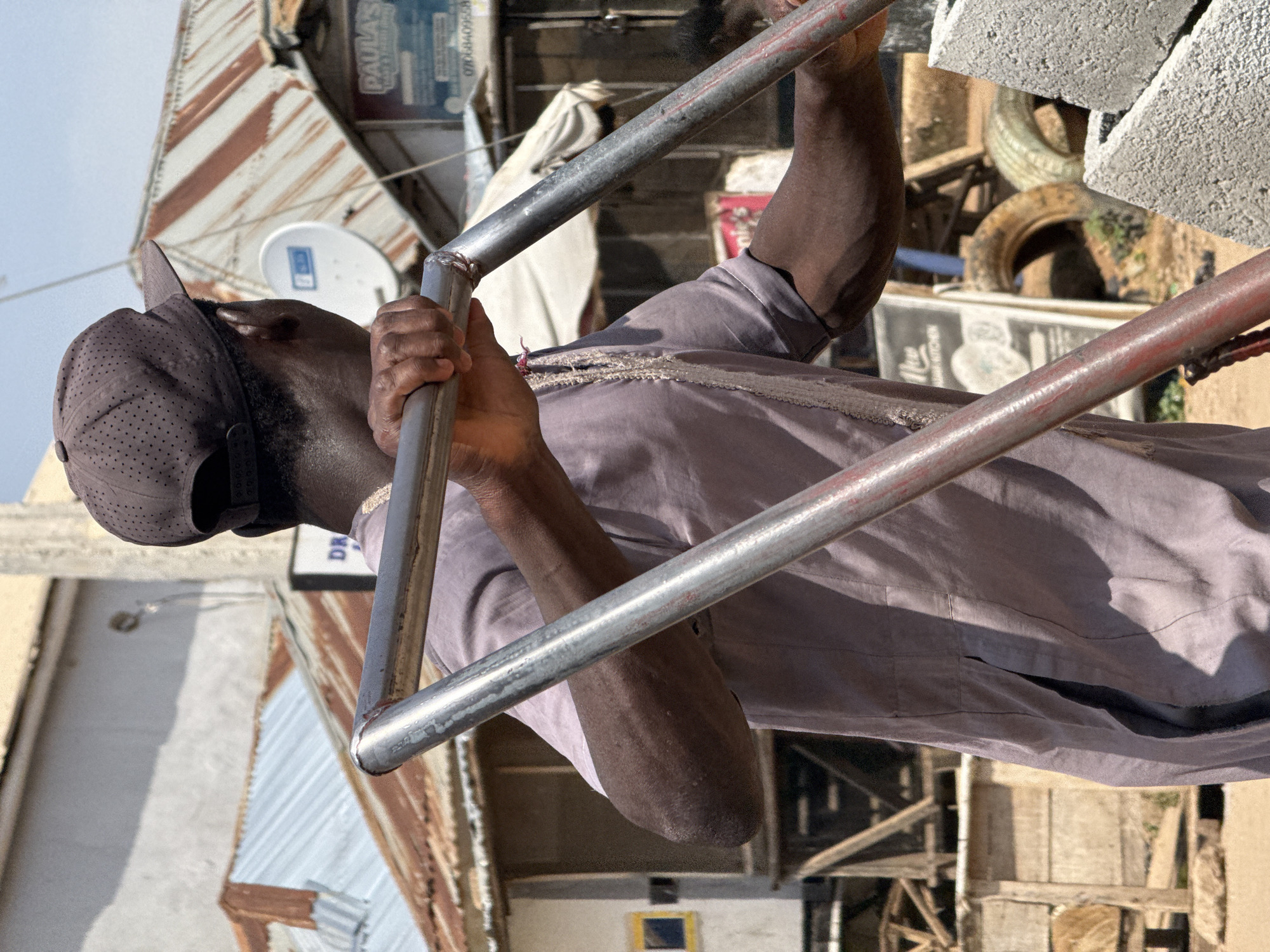 A close-up shot captures a Black man from the chest up, wearing a brown baseball cap and a light purple tunic, as he pushes a metal cart through a bustling urban environment. His hands grip the cart's handles, one hand visible on the left and the other on the right. The man is positioned in the foreground, with his back partially turned to the camera as he looks towards the right.
The background reveals a vibrant, somewhat disheveled street scene in Abuja, Nigeria. A collection of weathered buildings with corrugated metal roofs line the street, hinting at the lively, yet potentially informal, economic activity of the area. Signs and advertisements are visible on some of the buildings, including one for "Paula's" with a phone number that is partially legible. Scattered throughout the scene are various objects, such as tires, construction materials, and what appear to be pieces of wood, suggesting ongoing construction or repair work. The overall impression is one of a dynamic and active urban setting.