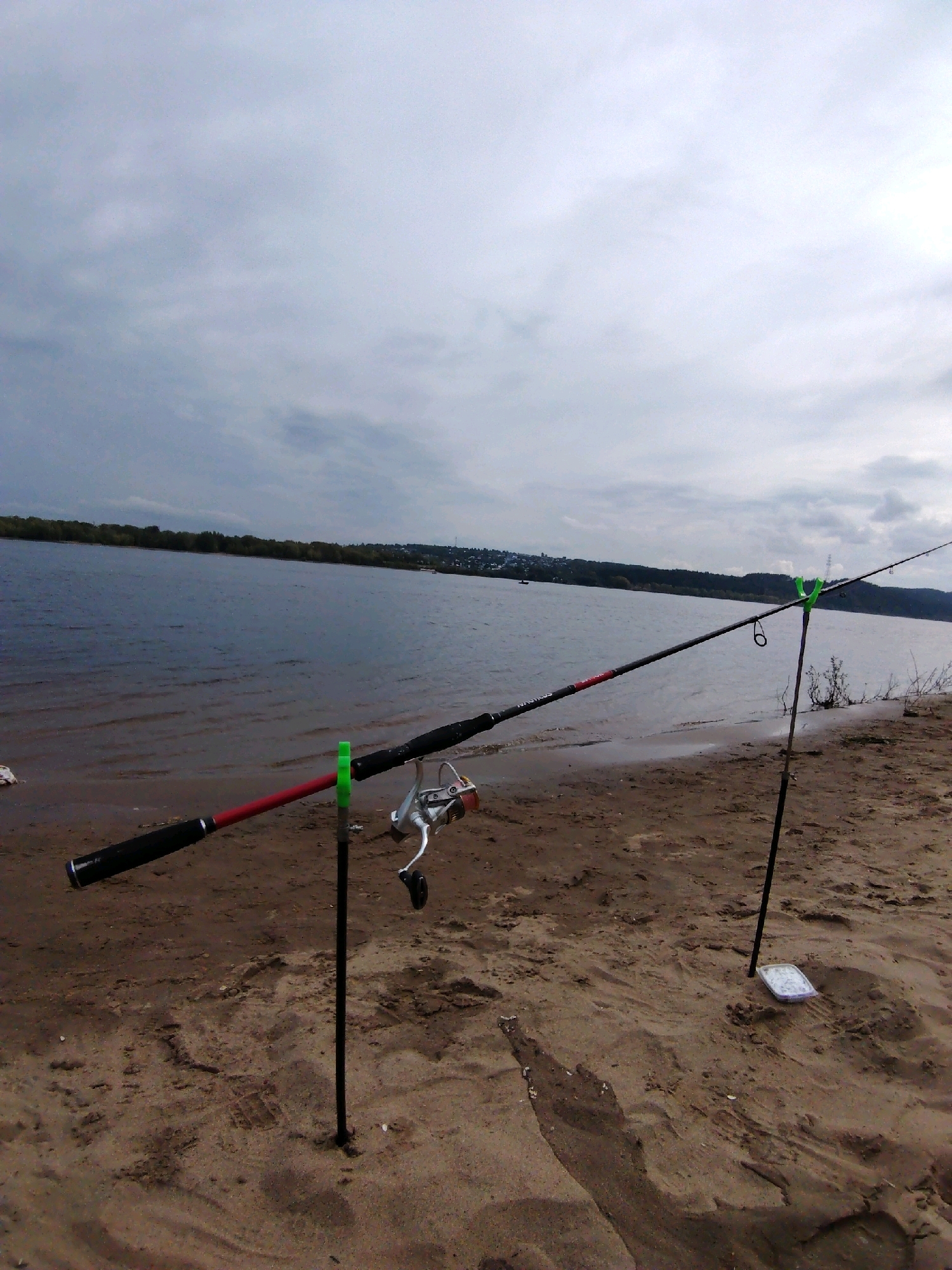 The image shows a sandy riverbank with two fishing rods set up on stands. The river is to the left, and the sky is overcast. The foreground is dominated by sand with footprints visible. One fishing rod, with a red accent, rests on a green rod holder. A fishing reel is attached to this rod. Another fishing rod is set up further back on a black stand with a green rod holder. A small white container is placed on the sand near the base of the second rod. The background shows a treeline and a hazy distant town. The scene is consistent with the Noviy, Russia location, likely near the Volga River, given the wide expanse of water and sandy banks.