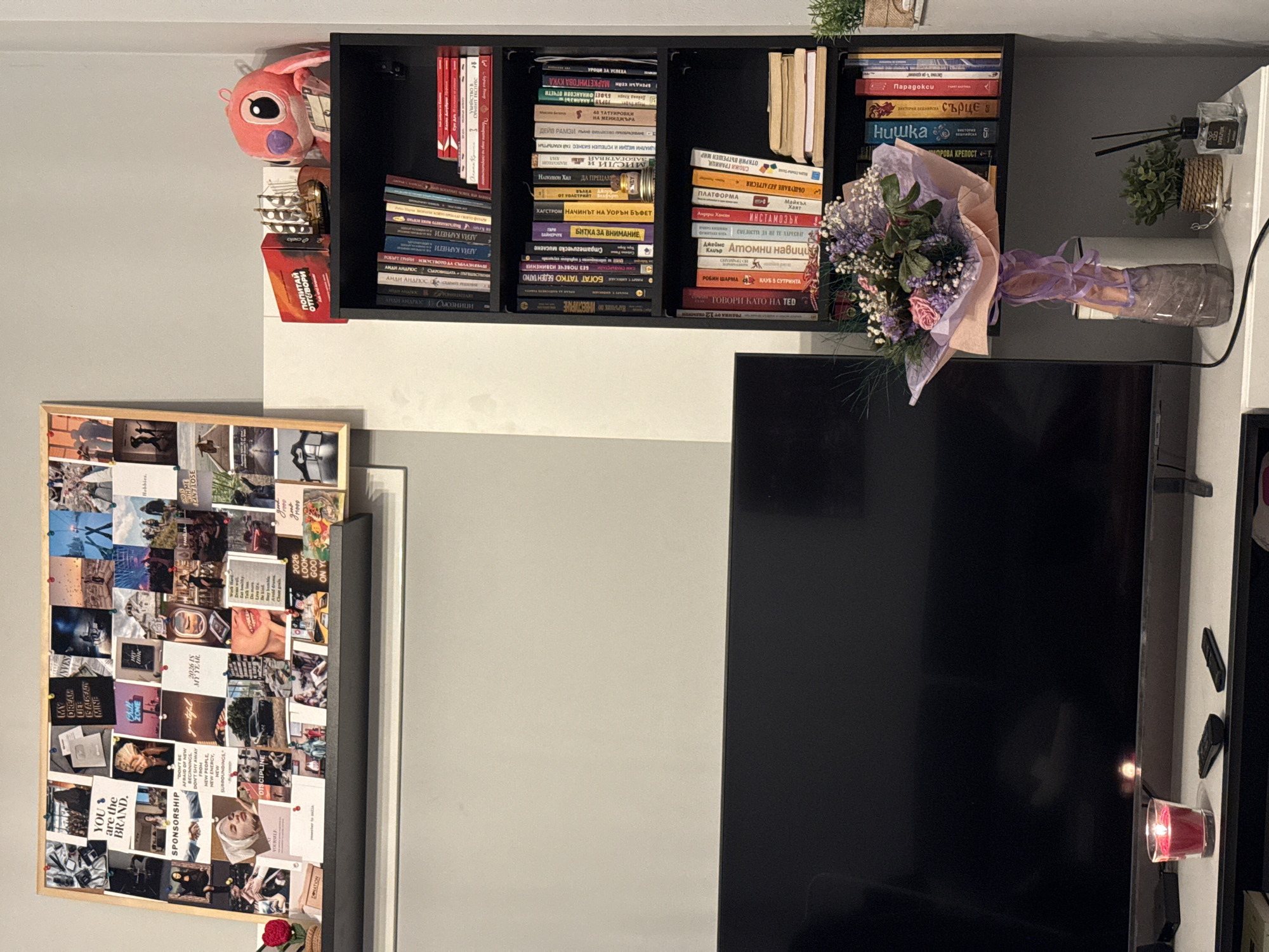 The image shows a section of a room, likely an apartment, in Sofia, Bulgaria, based on the Cyrillic script on the books. A black shelf unit is mounted on a gray and white wall, holding a variety of books, a pink stuffed animal, small decorative objects, and a bouquet of flowers in a light pink paper wrapping. Below the shelf is a flat-screen television. To the left of the television is a framed collage of photos and text on a wall, suggesting an inspiration board. A small red candle is on a ledge beside the TV. The room has a modern and decorated feel.