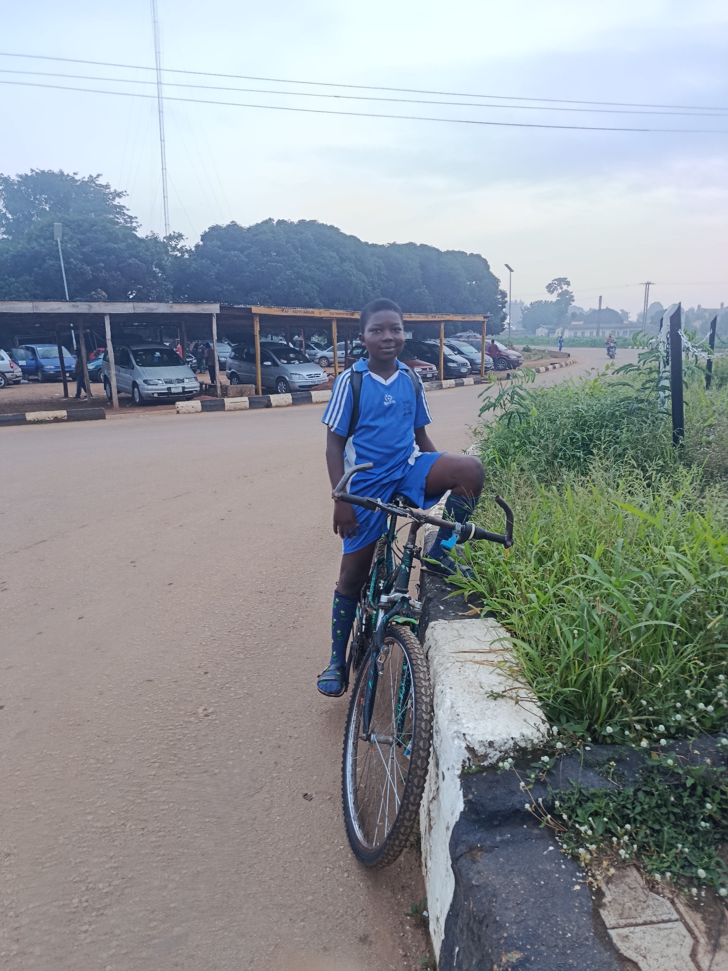 A young boy in a blue and white striped t-shirt and shorts, with green and white patterned socks, sits on a black bicycle with blue accents. He is positioned on a dirt road in front of a low concrete barrier overgrown with grass and small white flowers. The boy is looking at the camera with a slight smile. Behind him, a row of parked cars is visible under a covered parking area, with some people milling about. In the background, trees form a dense green canopy. Further in the distance, a tall radio tower stretches into a hazy sky, and power lines are visible. The overall scene suggests an outdoor environment in a town or city, possibly a public parking lot or roadside area in Maigizo, Nigeria, given the context provided. The lighting suggests it could be early morning or late afternoon.