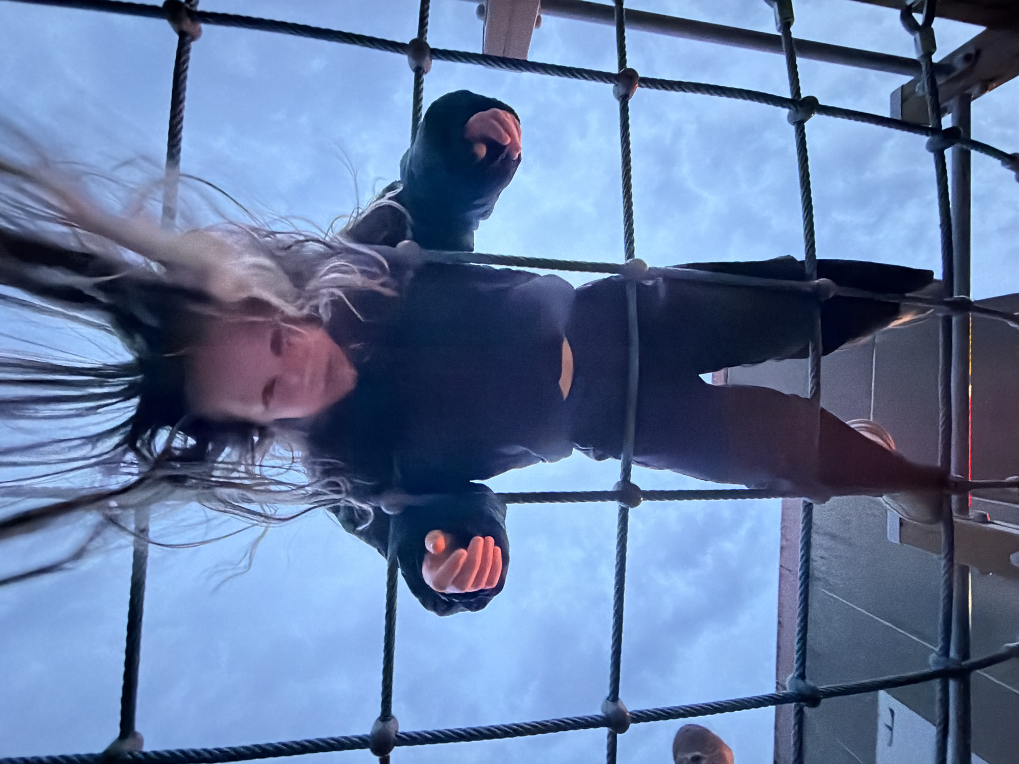 A person, likely a child, is upside down and suspended in a rope climbing structure, looking down at the camera. The subject is wearing dark clothing, including what appears to be a hooded jacket. Their hair is partially blonde and disheveled, hanging down due to the upside-down position. The rope structure is composed of thick, dark rope tied in a grid pattern. Beyond the rope, the sky is visible, appearing overcast. Parts of a building or playground structure are seen on the right side of the frame. There is no information to indicate this photo was taken in Марковский, Russia.