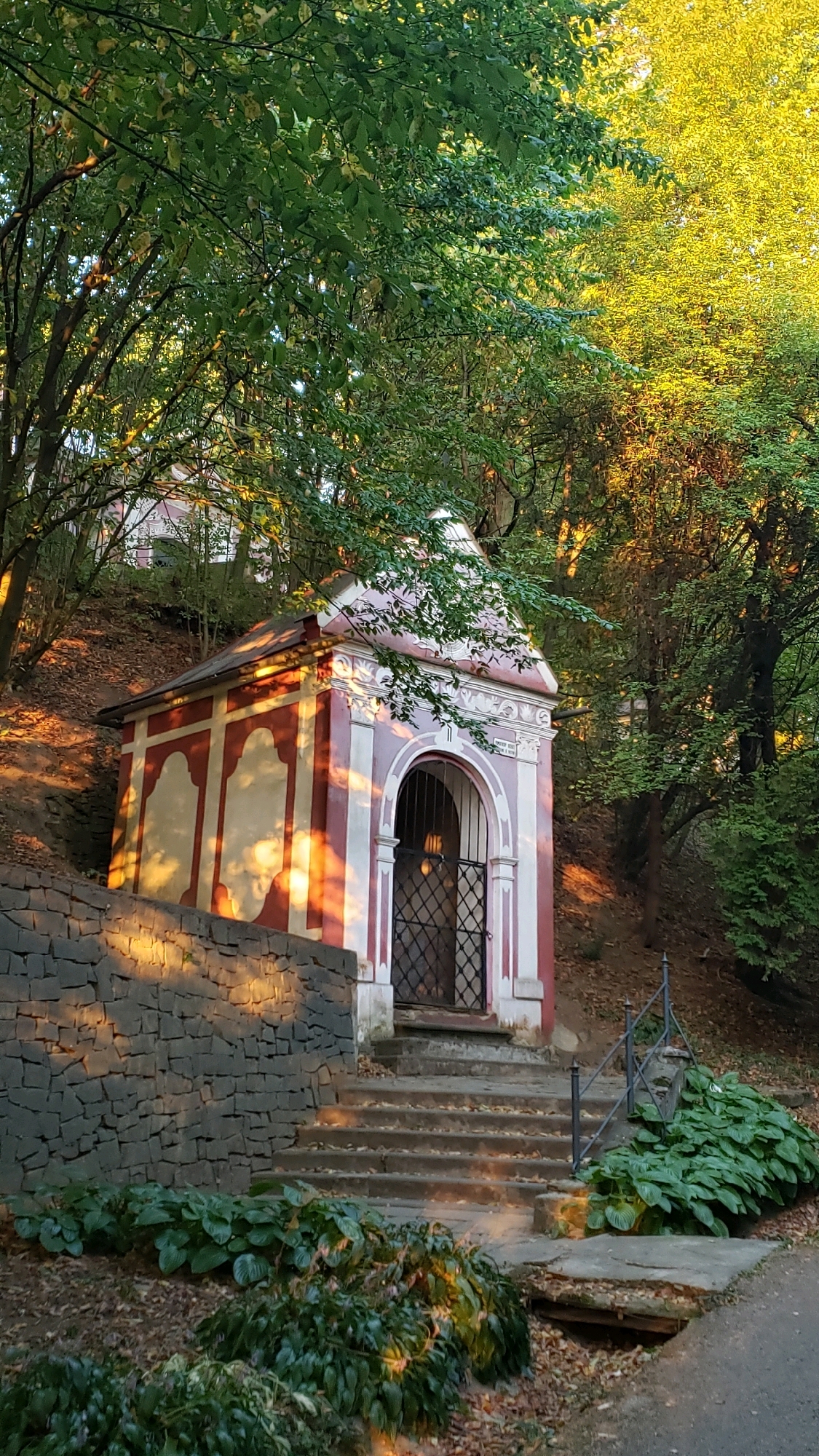 The image depicts a small, red and white chapel nestled amidst a wooded hillside. The chapel features an arched entrance with a wrought-iron gate. A stone wall runs along the left side of the scene, and a set of stone stairs leads up to the chapel entrance. Lush green foliage surrounds the chapel, with sunlight filtering through the trees, casting dappled shadows on the building and the surrounding ground. The scene appears to be in a natural setting, likely a park or a historical site. Based on available information, this chapel is likely located in the city of Prešov, Slovakia, specifically at the Kvačalova ulica viewpoint, also known as the Calvary viewpoint.