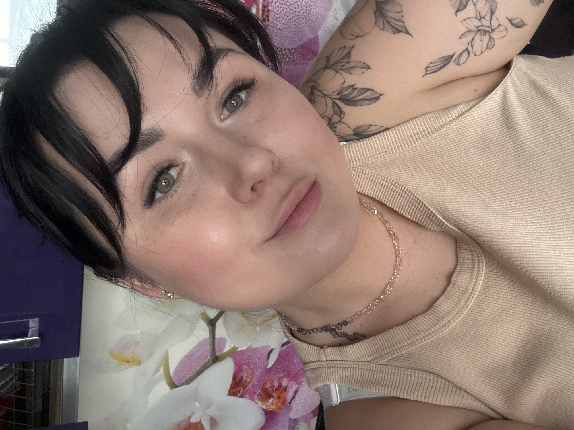 A medium close-up, slightly angled, portrait photograph depicts a light-skinned woman with dark hair framing her face. Her eyes are light, appearing grey-green, and her lips are lightly colored and pursed. A scattering of freckles is visible on her cheeks.
She wears a beige ribbed tank top. A delicate, layered necklace adorns her neck. A tattoo of floral and leaf patterns is visible on her upper arm and shoulder. Small earrings are present in her ear.
Behind her, a floral patterned surface with white and purple orchids is partially visible. A dark purple object, possibly a cabinet, is also present in the background.
The location is not discernible as Chaikovsky, Russia based solely on the visual information provided in the photograph.