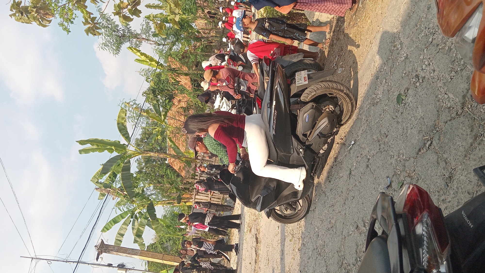 In the city of Kedungwaru, Indonesia, a woman with long dark hair, wearing a maroon off-the-shoulder top and white flared pants, rides a black scooter. She is in the foreground, facing left and looking forward. Behind her, a man with a brown cap and a green patterned shirt is also on a scooter, facing left. Several other people, including children, are visible in the background, some wearing red shirts and hats, others in dark clothing. The environment appears to be an outdoor, dusty area with trees, including banana trees, and buildings with thatched roofs. Power lines are visible overhead, and a concrete pillar stands on the left side of the frame. The overall impression is a sunny day with a gathering or event taking place.