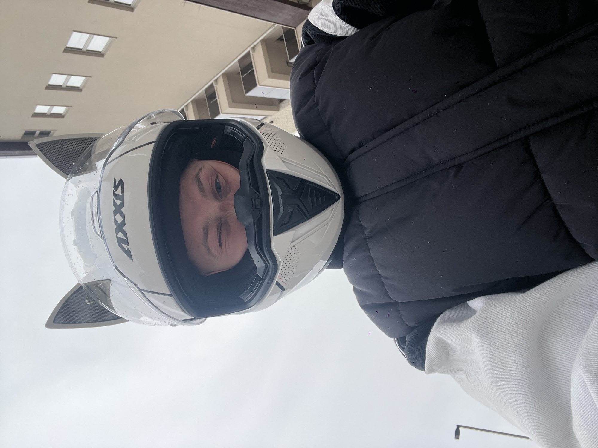 A person wearing a white motorcycle helmet is pictured, facing the camera. The helmet has clear rain drops on the visor and decorative cat ears. The person also wears a puffy black jacket with a white hoodie underneath. In the background are light-colored buildings with windows, indicative of urban architecture. The setting is suggested to be the city of Bečići, Montenegro. The image is oriented in a rotated perspective.