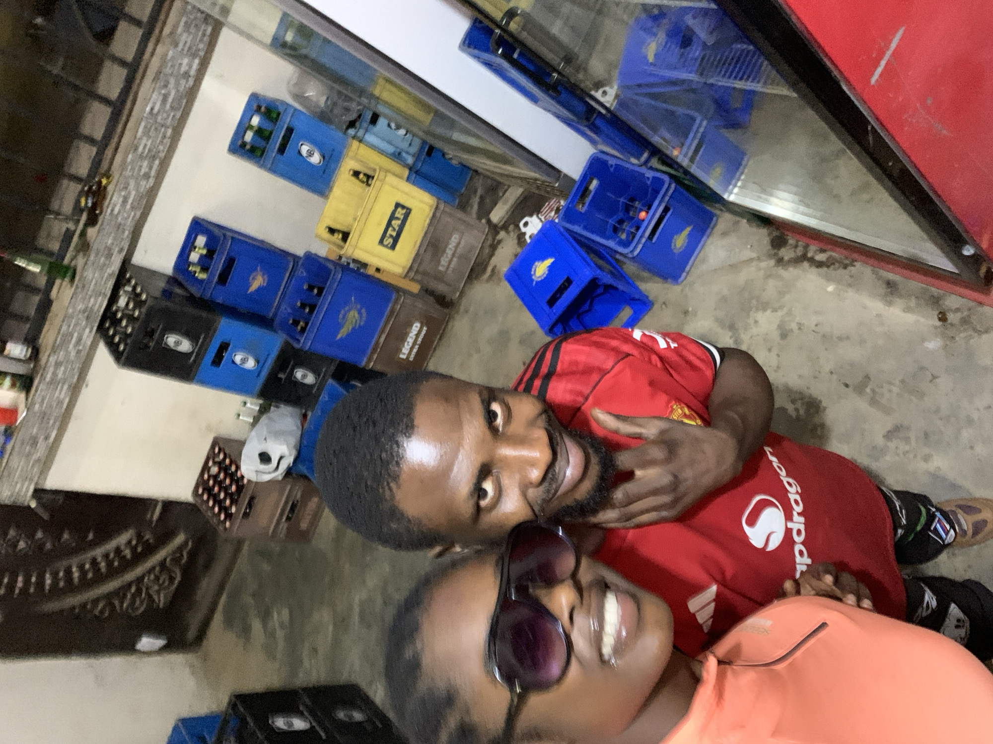 The image is an upward-angled selfie of a man and a woman. The man is wearing a red Manchester United soccer jersey with the Snapdragon logo and has a beard. The woman is wearing sunglasses and a peach-colored shirt. Behind them are stacked crates of beer bottles of various brands (Star, Legend, etc.) and other containers and storage items. It is implied that the location is near the New Stadium in Nigeria.