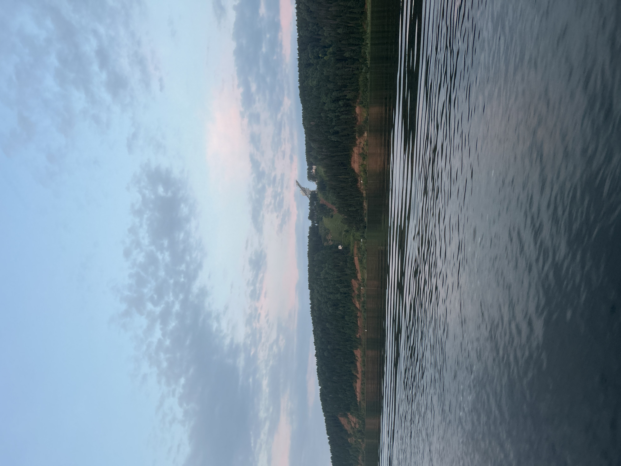 This is a photograph of the Volga River near Noviy, Russia, taken from a boat. The sky is a pale blue with scattered clouds, tinged with pink and lavender from the setting sun. A dense, dark green forest covers the steep riverbank on the far side of the water. On the crest of the hill, a modern ski jump structure is visible, with a small building nearby. The water in the foreground is dark and rippled, reflecting the muted colors of the sky. There are no people or animals visible in the frame.