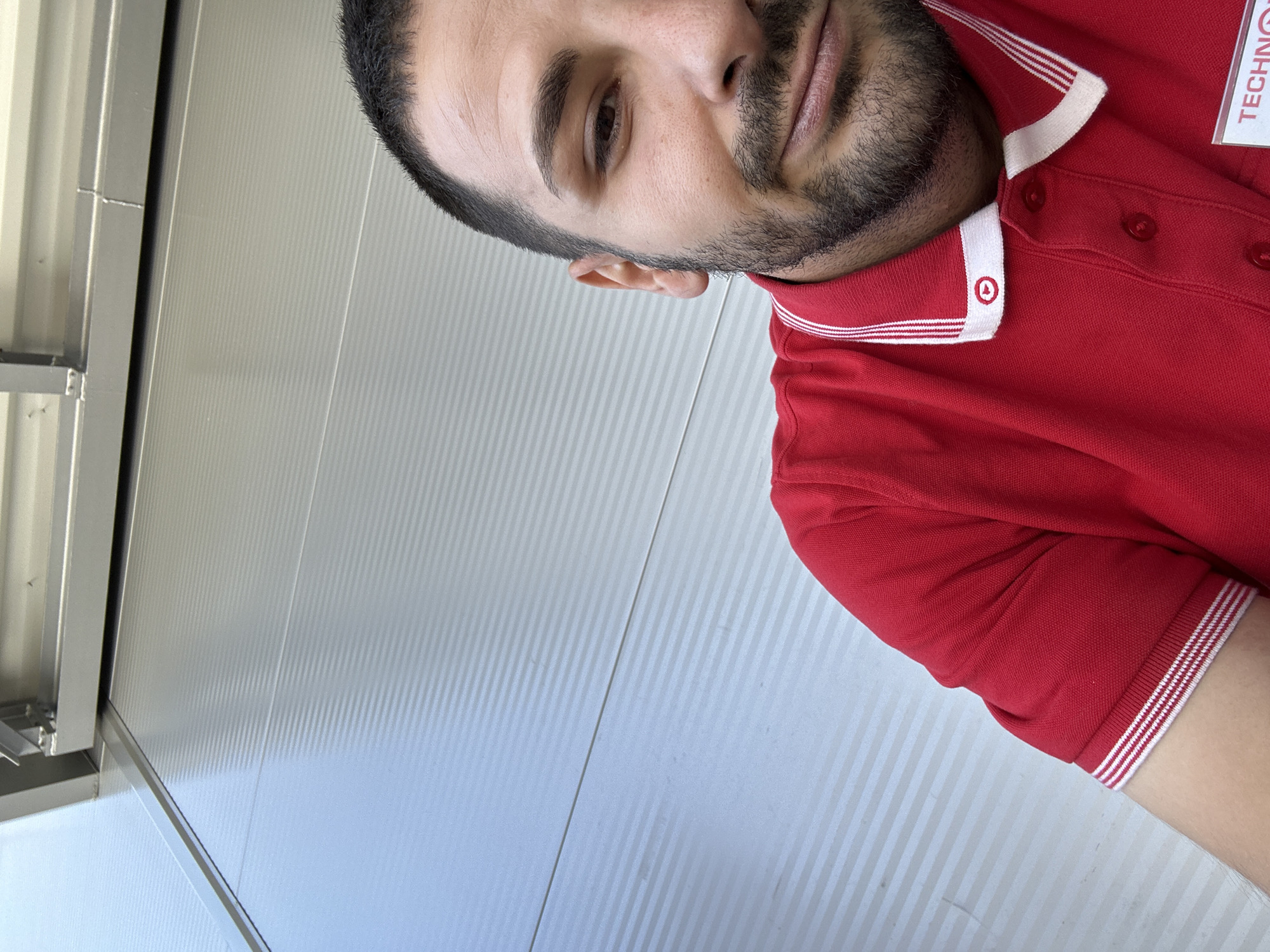 The image shows a man with dark hair and a beard wearing a red polo shirt with white stripes on the collar and sleeves. The shirt has buttons and a logo on the collar. The background consists of a corrugated metal wall with horizontal lines. The location is Unknown, Unknown.