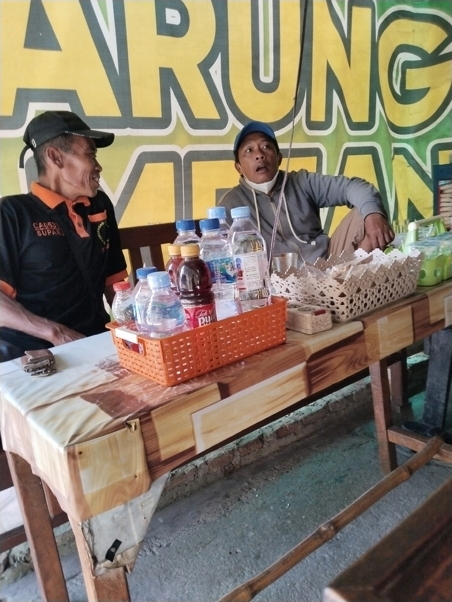 The image depicts a roadside food stall in Trenggalek, Indonesia. Two men are seated at a wooden table covered with a patterned tablecloth. The man on the left, wearing a black polo shirt with orange trim and a black baseball cap, is looking to the left and smiling. The man on the right, wearing a gray hooded sweatshirt and a blue baseball cap, is looking forward with his mouth open, as if speaking.
On the table are several bottles of water and tea, arranged in an orange plastic basket. To the right, there's a white woven basket containing what appear to be snacks and condiments. The background features a large yellow and green banner with text that is partially visible but likely advertises the stall's offerings. The overall environment suggests a casual, open-air setting typical of local food vendors in Indonesia.