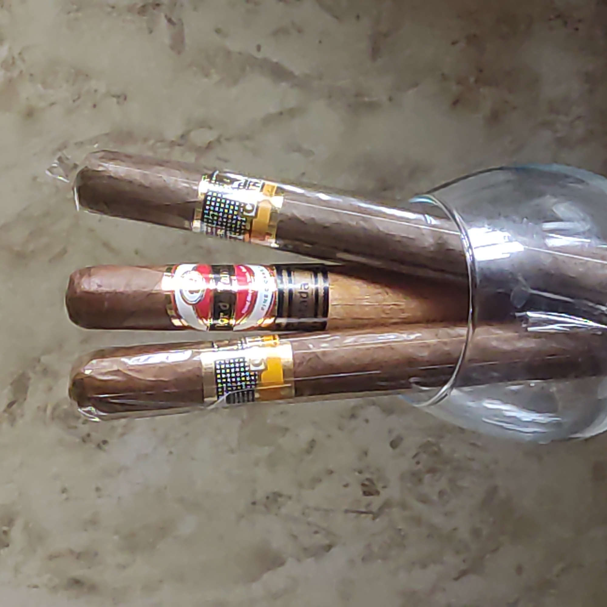 Three cigars, individually cellophane-wrapped, stand upright in a clear glass bowl. The cigars are a medium brown color with visible tobacco leaf texture. Each cigar has a band around it, with the center cigar's band featuring red, white, and gold detailing. The background is a mottled, textured surface that appears to be a wall or countertop with variations in color and pattern. The lighting is somewhat diffused, creating soft shadows. There are no people present in the image, and the location is not discernible from the visual information provided.