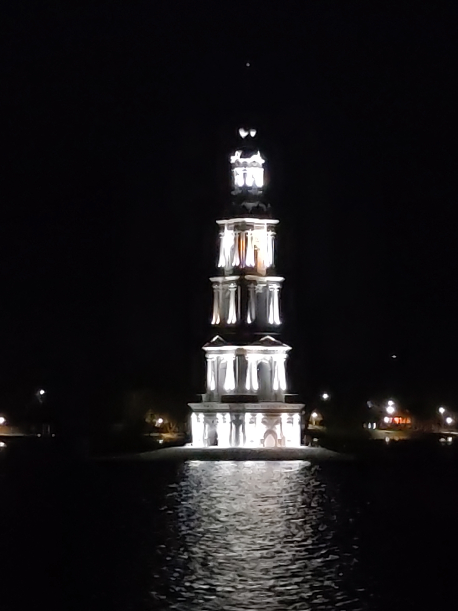 Captured at night, the scene portrays the Kalyazin Bell Tower situated in the city of Kalyazin, Russia. The tower, illuminated with bright lights, rises from the water, reflecting its glow on the water's surface. The structure has a tiered design and is of a light hue. The surrounding environment is dark, punctuated by a few lights on the horizon. The atmosphere has a serene, ethereal quality.