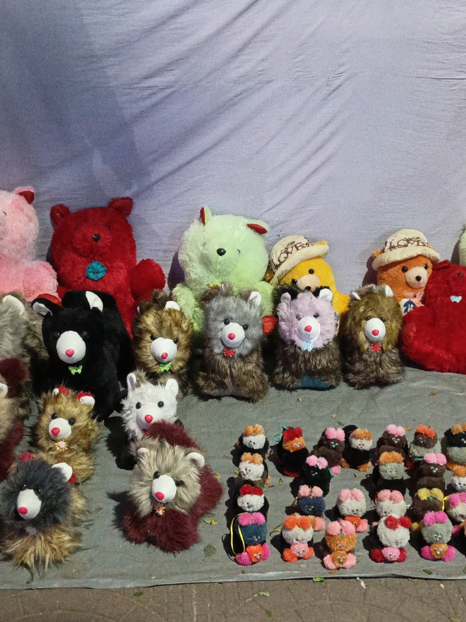 The image shows a collection of teddy bears of various sizes, colors, and styles arranged on a gray surface, likely a cloth or mat, against a light-colored backdrop. There are pink, red, green, yellow, and orange bears in the background, along with smaller, furry teddy bears with different colored collars. In the foreground, there are several smaller teddy bears with fluffy bodies and differently colored details. Some of the bears have hats and bows. At the lower right corner, there is a collection of tiny pom-pom bears. The setting suggests an outdoor market or a display of toys, possibly in Dhaka, Bangladesh. There are no people directly visible in the image.