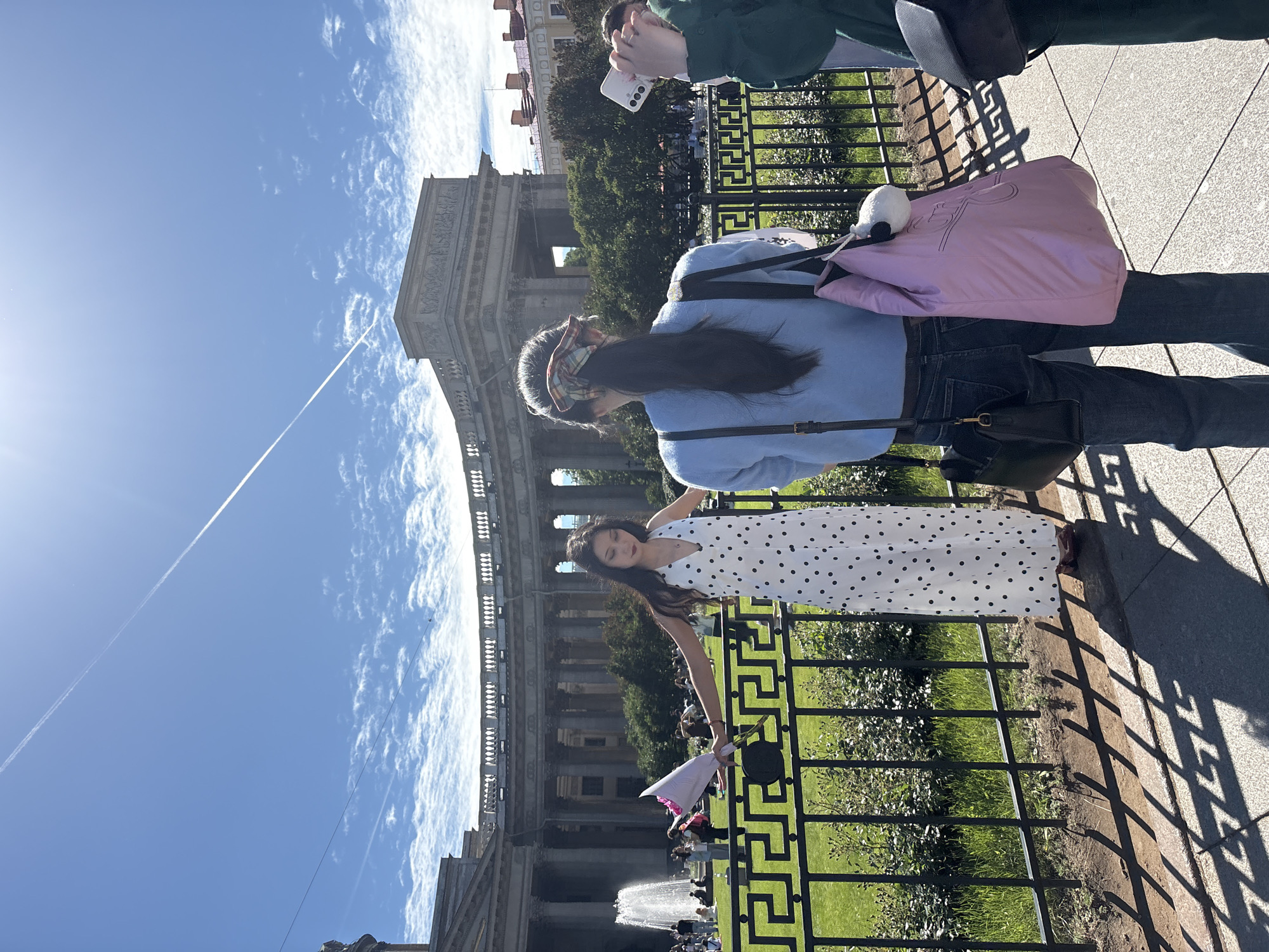 The image captures a sunny day in Saint Petersburg, Russia, likely near the Kazan Cathedral, a prominent architectural landmark. In the foreground, a young woman stands to the left, wearing a long white dress with black polka dots and holding a bouquet of pink flowers. Her arms are outstretched, and she is looking downwards. Behind her, another person is seen from the back, wearing a blue sweater and jeans, with a pink bag slung over their shoulder. This individual is holding a smartphone, possibly taking a photo. The background features the grand facade of the Kazan Cathedral with its colonnades and a fountain. Several other people are visible in the park area behind the fence, enjoying the day. The sky is blue with scattered white clouds and a contrail from an airplane. The lighting suggests it is mid-day.