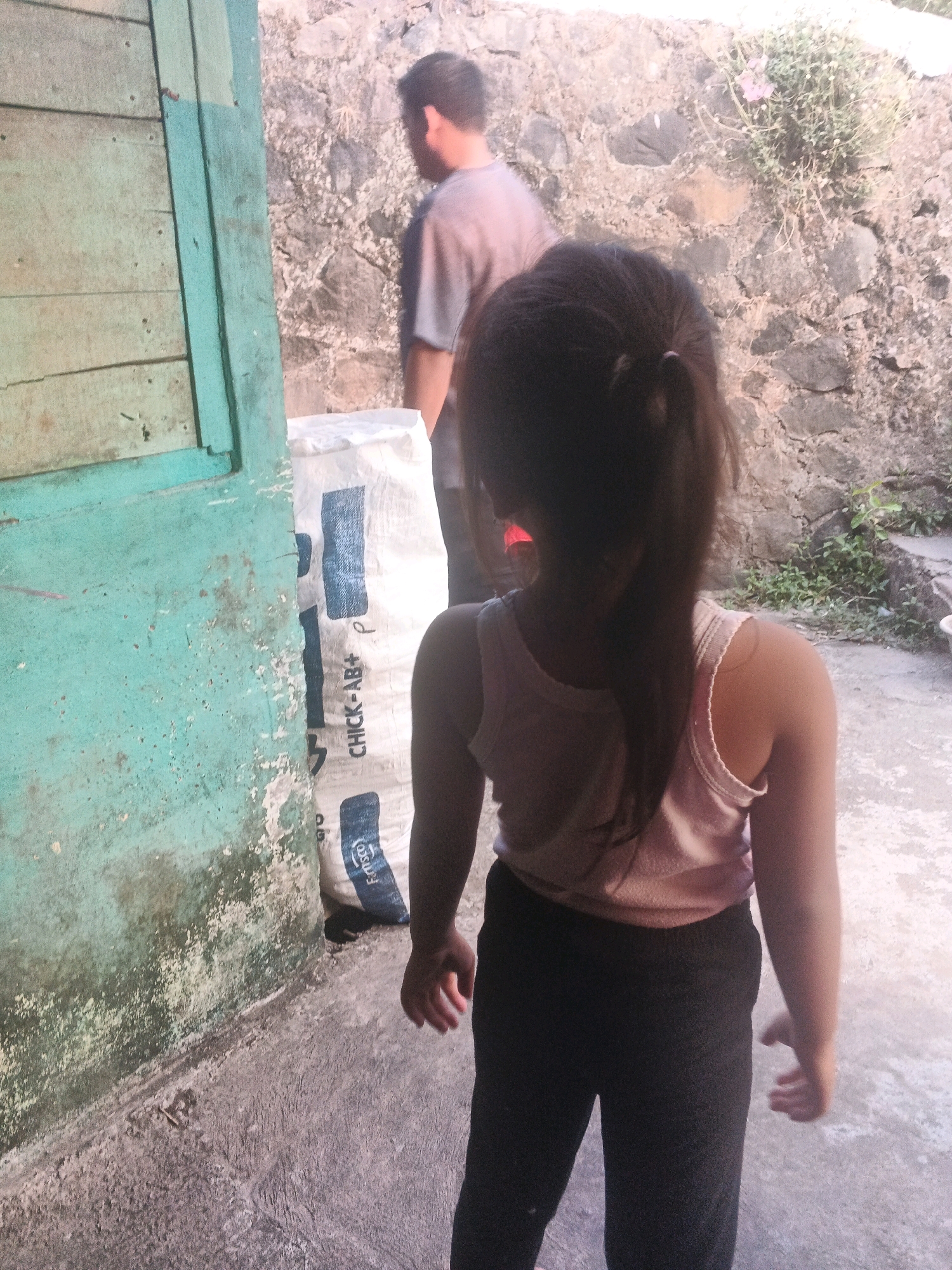 The image depicts a scene outdoors, likely in the city of Keji, Indonesia, with a focus on two individuals and their surroundings. In the foreground, a young girl is seen from behind, facing away from the camera. She has dark hair, which is pulled back, and is wearing a pink sleeveless top and black pants. Her right arm is extended slightly. To the left of the girl, a large white bag with blue markings is visible. Behind the girl, further in the background, a man is walking away from the camera. He is wearing a gray t-shirt. The environment appears somewhat rustic, with a textured stone wall behind the individuals and a weathered turquoise-colored structure to the far left of the frame. The ground is concrete.